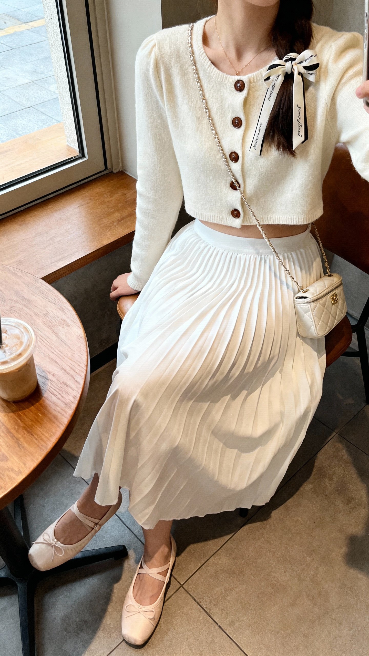 iPhone seated café selfie cropped at neck showing a woman in a white pleated skirt with a cropped cardigan buttoned at top, ballet flats, ribbon hair tie, and a tiny shoulder bag, face not visible, morning window light, natural iPhone photo quality.