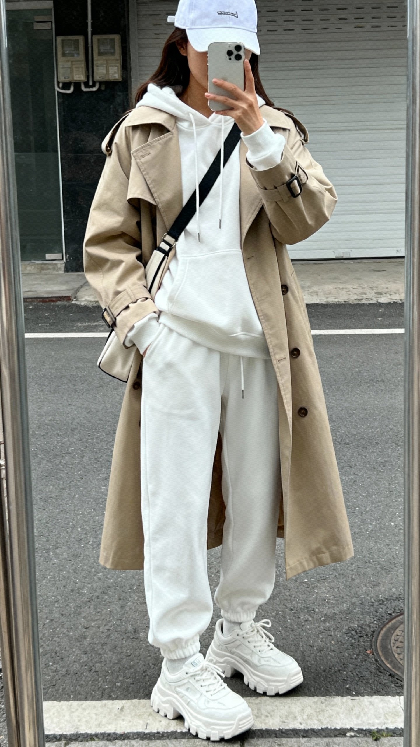 iPhone street-side mirror reflection selfie of a woman in a monochrome white hoodie under a beige trench, white joggers, chunky white sneakers, baseball cap, crossbody bag, face not visible, overcast natural light, casual iPhone photo.