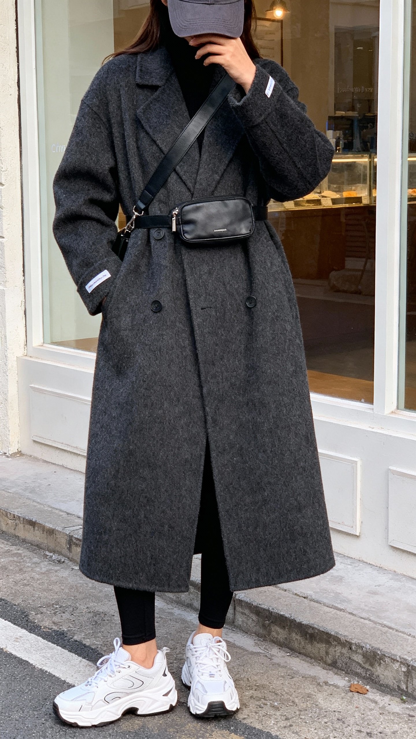 iPhone street-side selfie cropped at neck showing a longline charcoal wool coat, black leggings, chunky white sneakers, baseball cap, crossbody belt bag, face not visible, morning daylight by storefront window, natural iPhone quality.