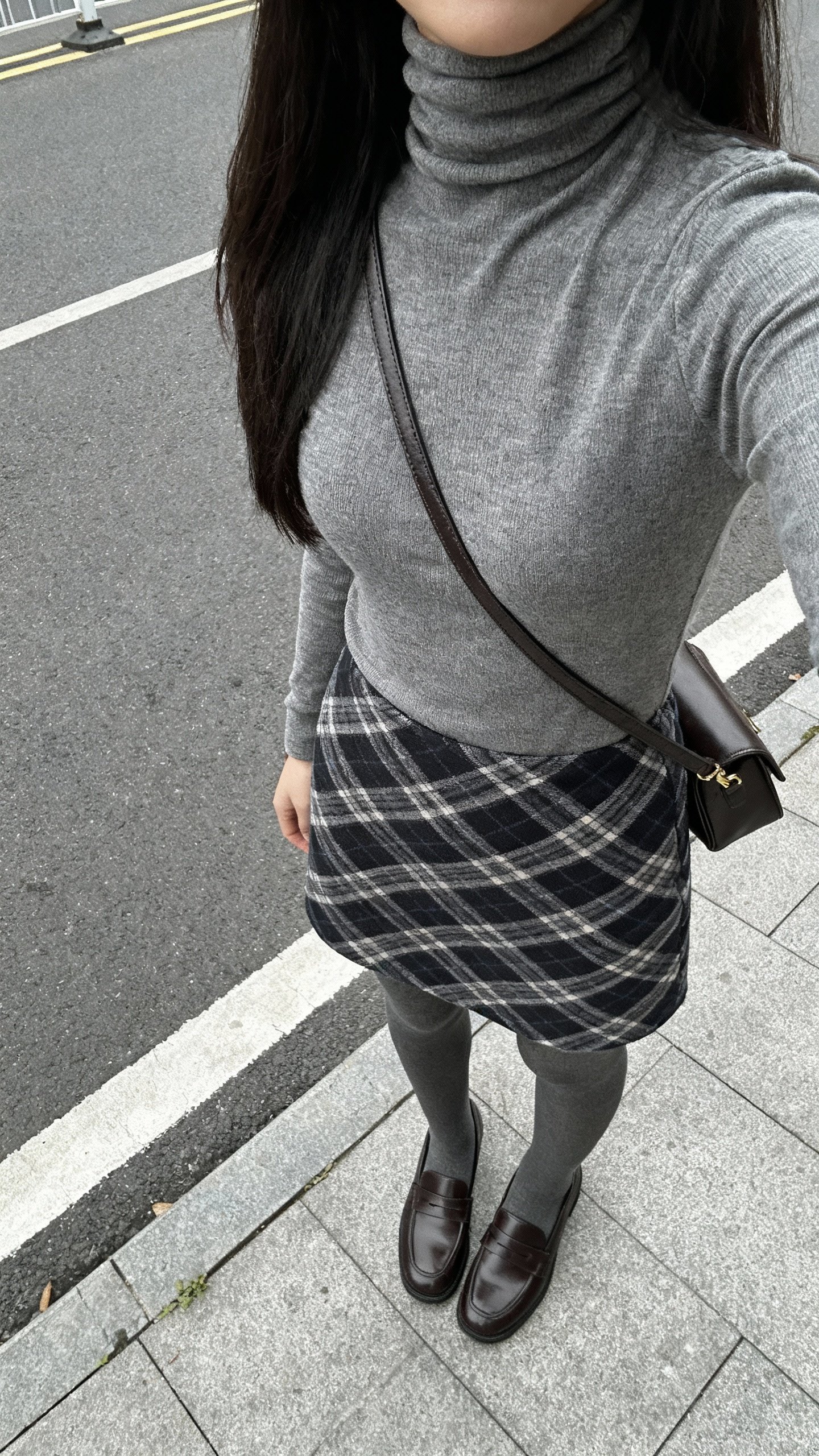 iPhone street-side selfie from the side angle showing a modern plaid skirt with a fitted turtleneck, opaque tights, loafers, crossbody bag, face not visible, overcast daylight, casual iPhone photo.