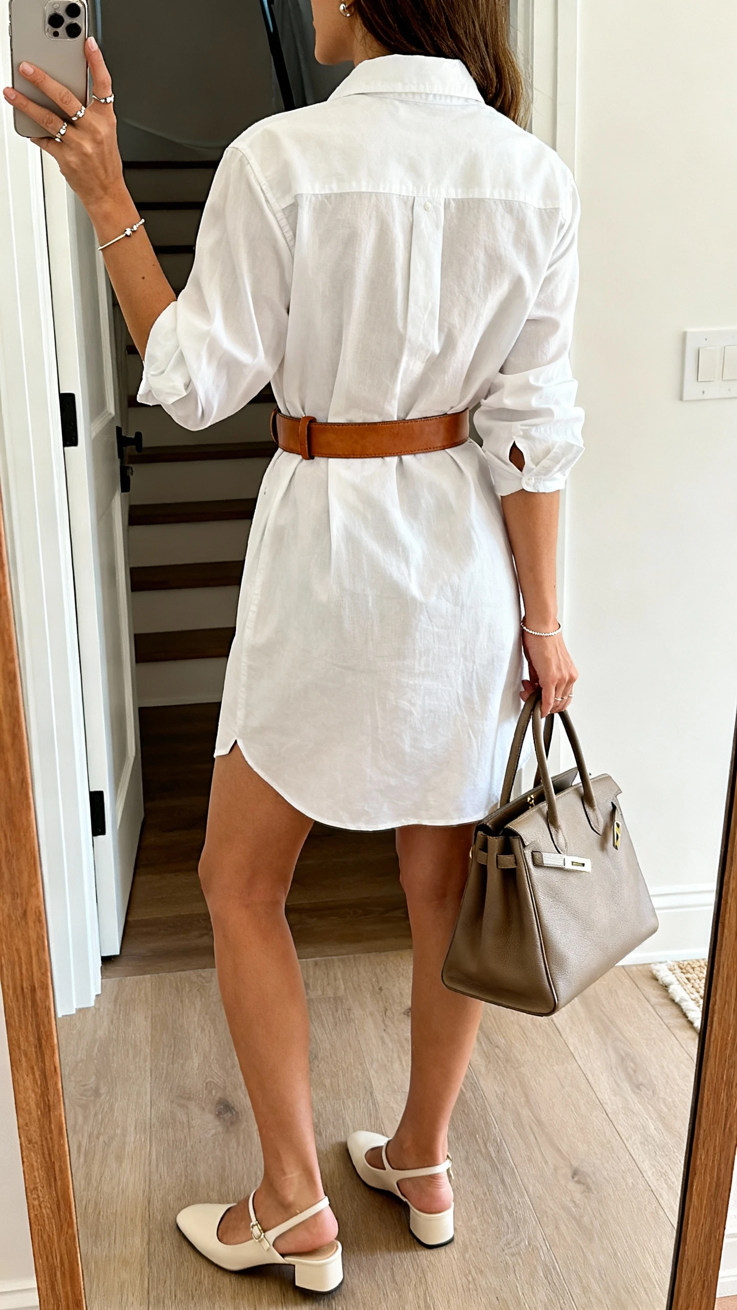 iPhone back-view selfie in a full-length mirror showing a woman in a crisp cotton poplin shirt dress cinched with a medium leather belt, clean slingbacks, simple studs and a dainty bracelet, polished tote in hand; entryway setting, daylight, face not visible, casual iPhone aesthetic.