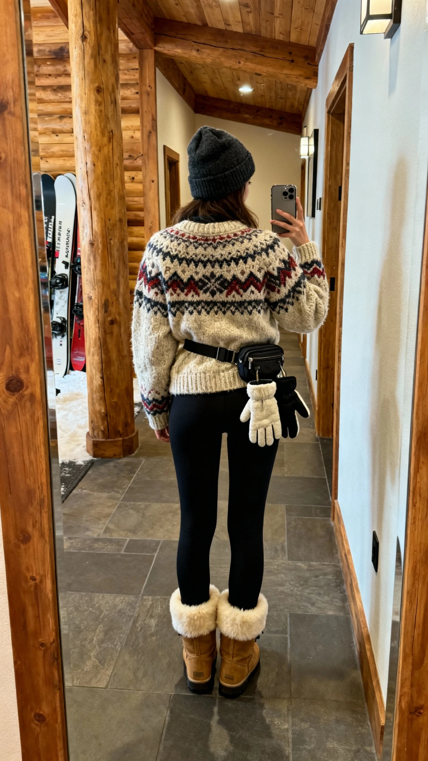 iPhone back-view selfie of a woman in a chunky Fair Isle knit sweater, fleece-lined black leggings, shearling-lined boots, beanie, mittens tucked in belt bag, face not visible, ski lodge hallway mirror, warm indoor lighting, natural iPhone photo quality.