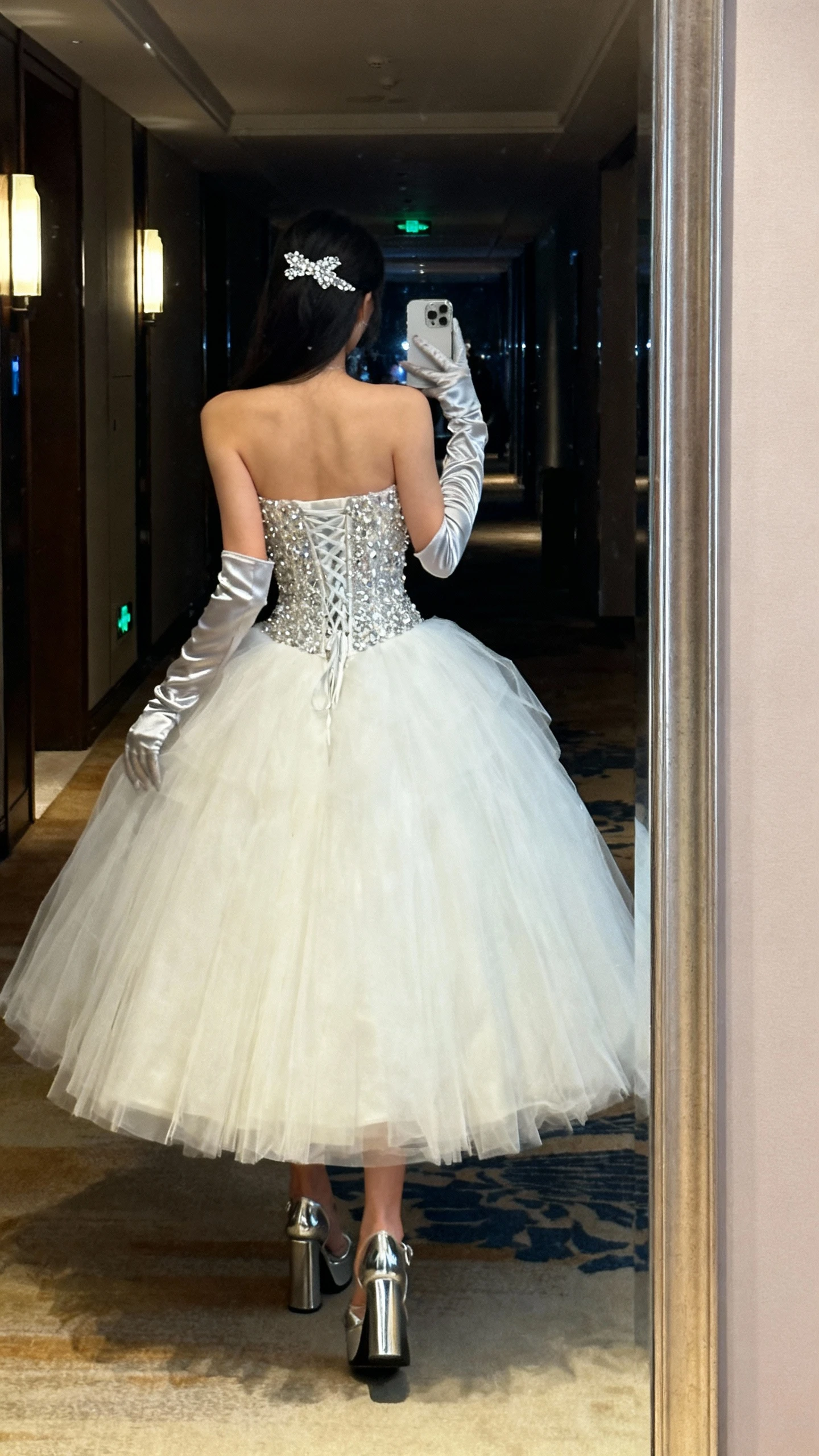iPhone back-view selfie of a woman in a crystal-corseted ballgown that owns the spotlight: voluminous tulle skirt, sparkling crystal corset top, satin gloves, metallic platform heels, crystal hair clip; face not visible, hallway mirror outside a gala venue, evening indoor lighting, natural iPhone photo.