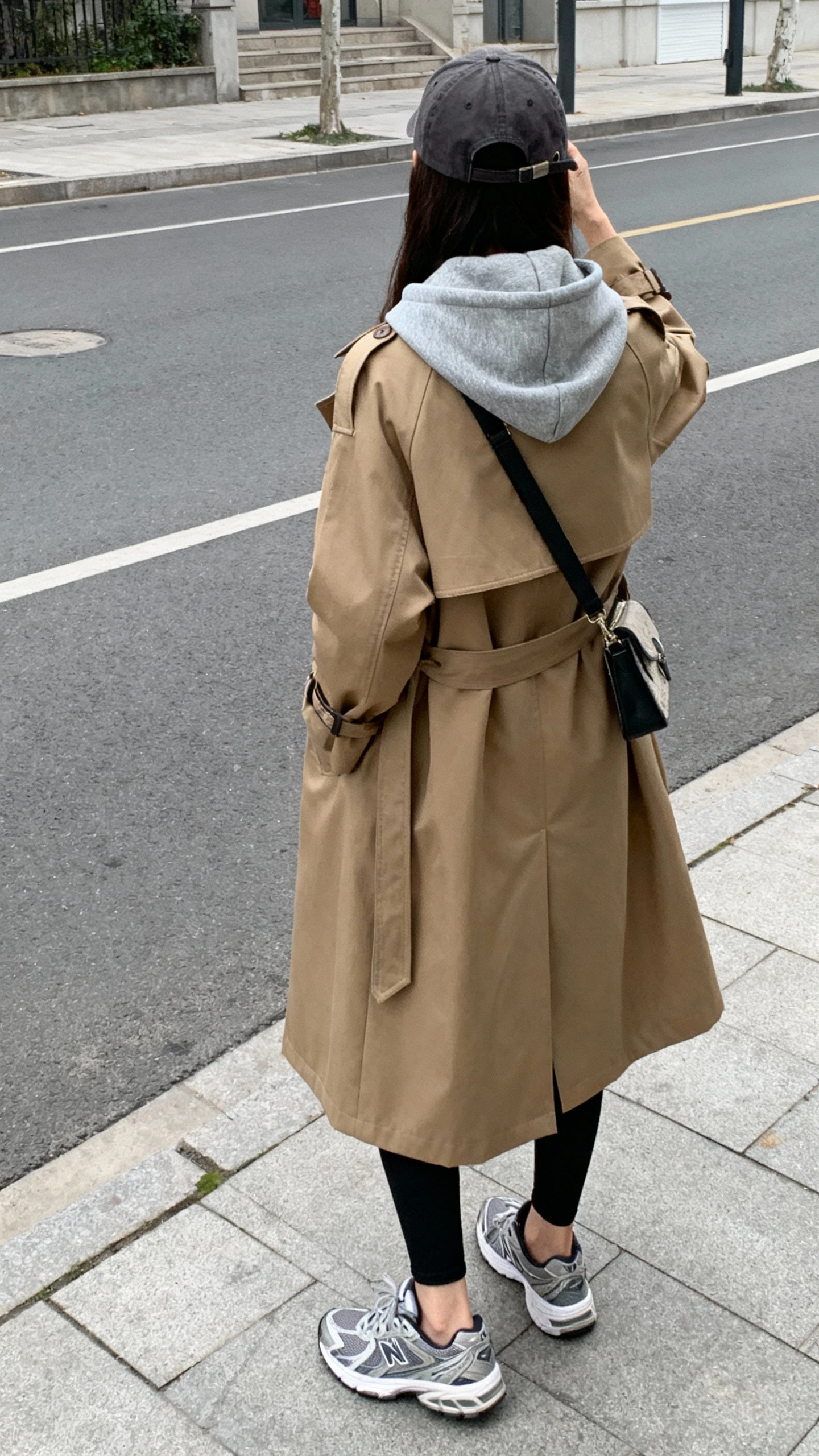 iPhone back-view selfie of a woman in a gray hoodie layered under a camel trench coat, black leggings, sporty sneakers, baseball cap, crossbody bag, face not visible, city street sidewalk after class, overcast daylight, natural iPhone photo.