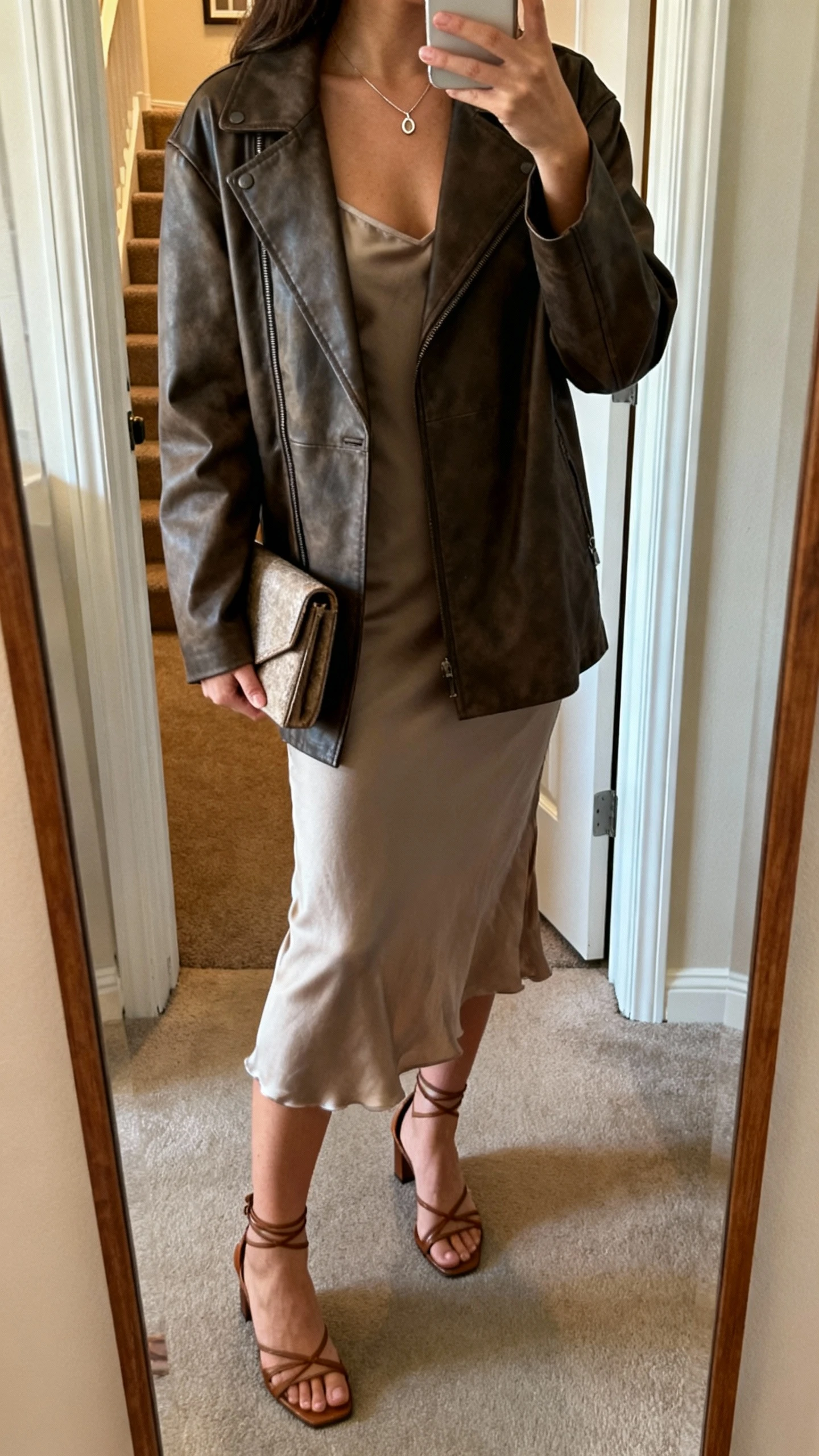 iPhone back-view selfie of a woman in a leather (or faux) jacket over a slip dress with strappy heels and a delicate pendant, clutch in hand, face not visible, apartment entry mirror, warm indoor light, natural iPhone quality.
