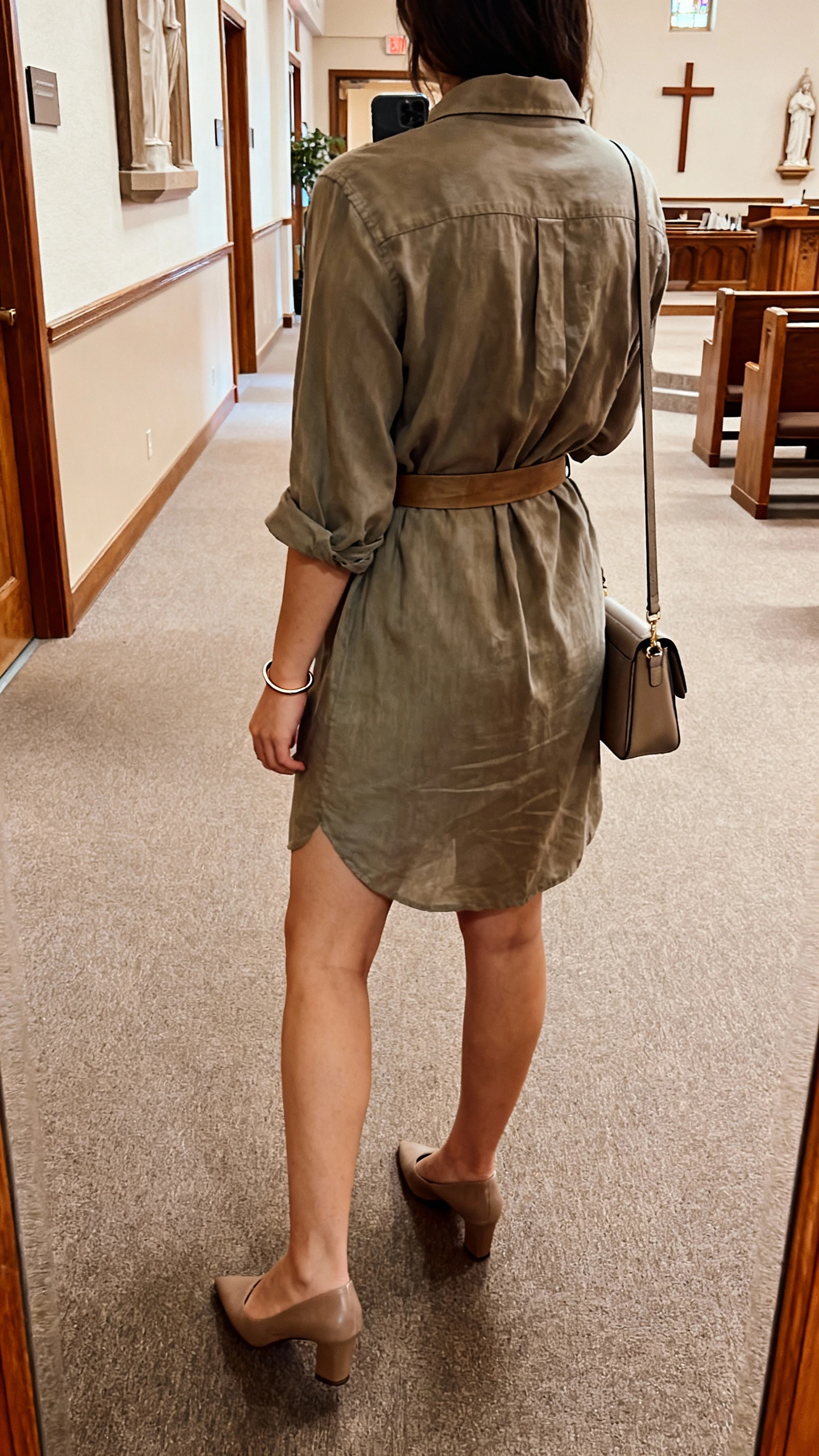 iPhone back-view selfie of a woman in a relaxed shirt dress cinched with a belt, low heels, slim bracelet, small shoulder bag, face not visible, church lobby hallway lighting, natural iPhone quality.
