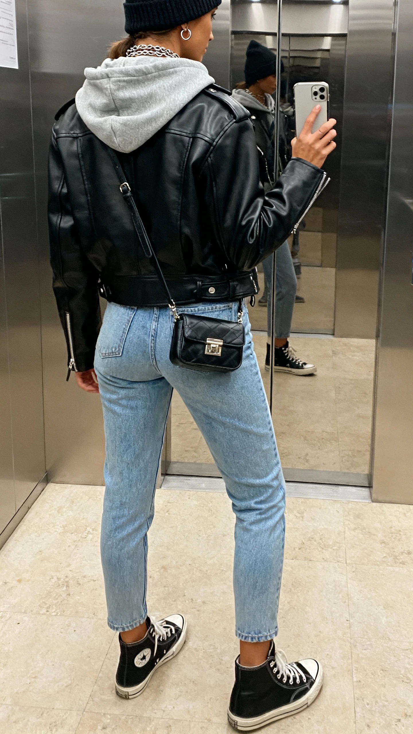 iPhone back-view selfie of a woman wearing a black leather biker jacket over a gray hoodie, light blue tapered jeans, black high-top sneakers, beanie, mini crossbody, layered chains and small hoops, face not visible, elevator mirror, indoor lighting, natural iPhone photo quality.