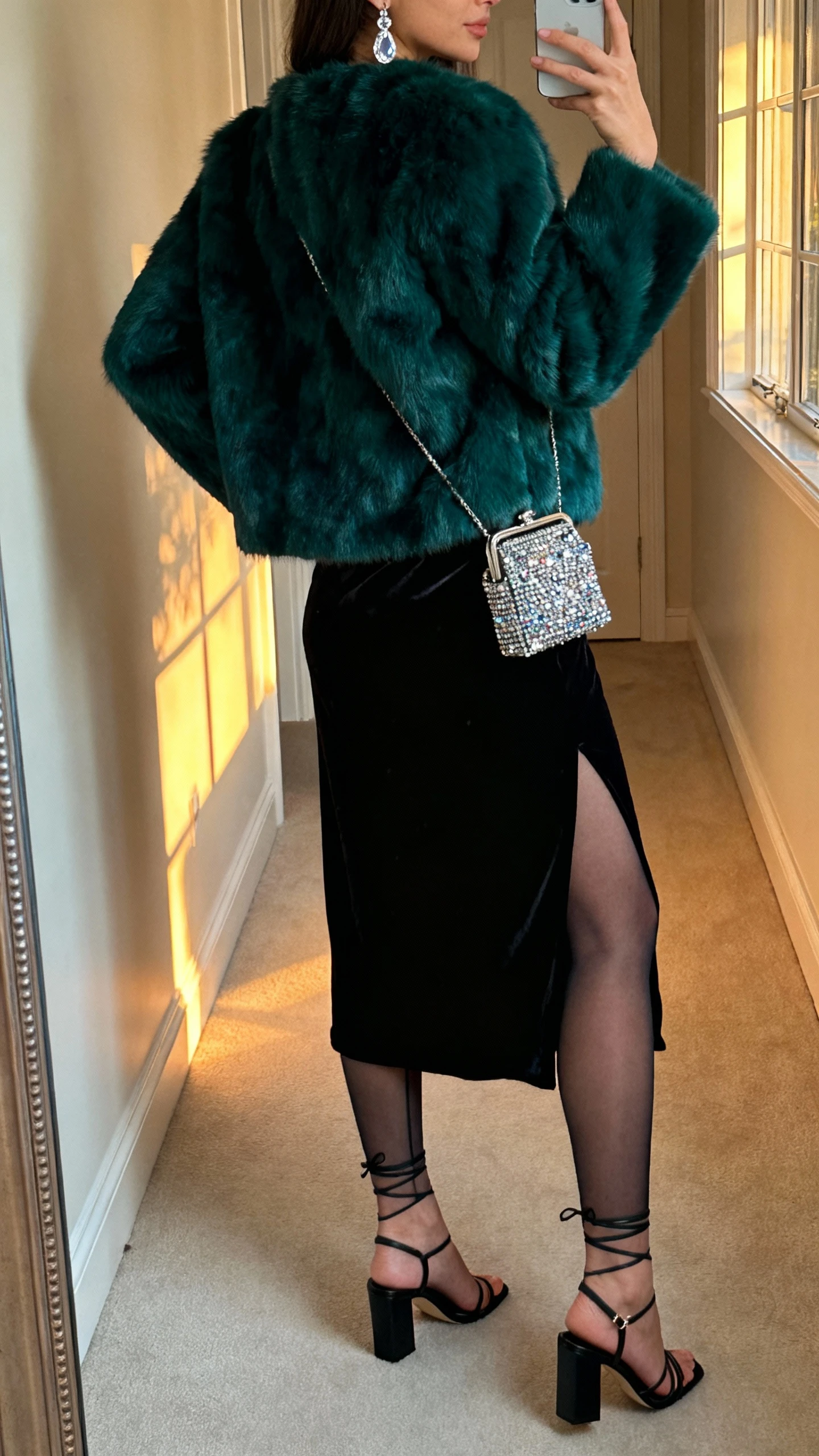 iPhone back-view selfie of a woman wearing a deep emerald faux fur jacket over a black velvet slip dress, sheer tights, strappy block-heel sandals, micro embellished mini bag, crystal drop earrings; hallway mirror, face not visible, evening indoor lighting with some window glow, natural iPhone photo quality.
