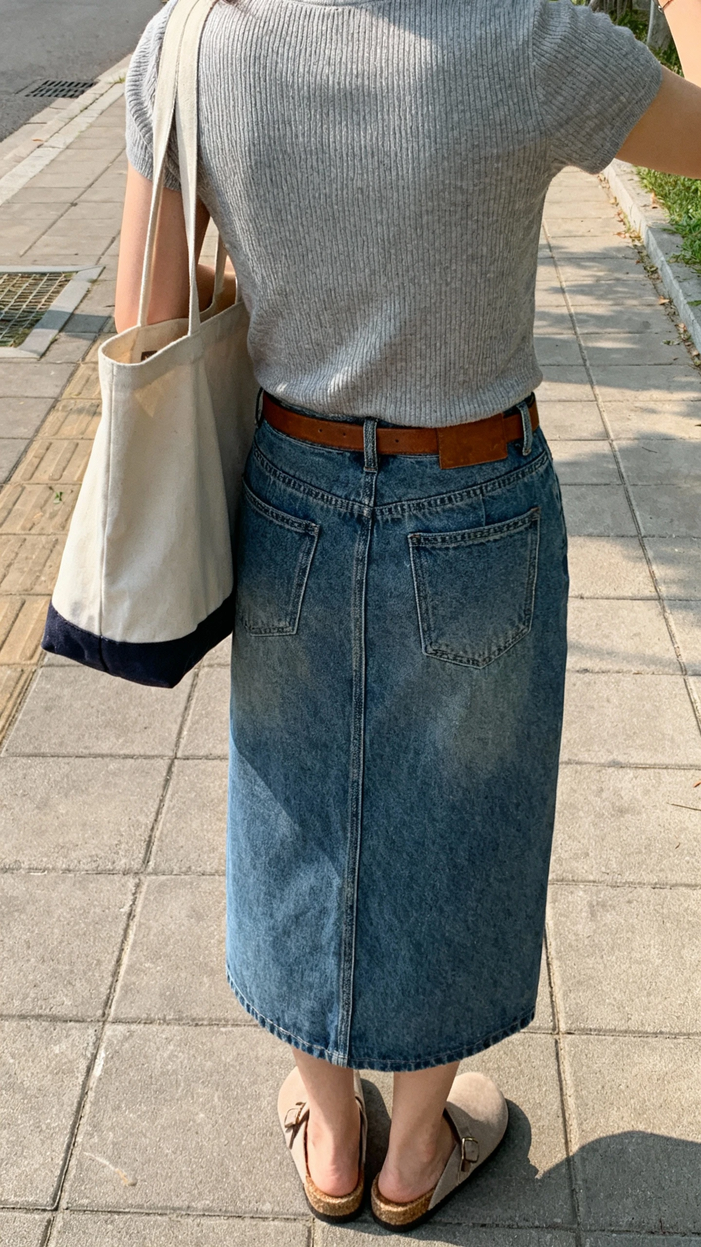 iPhone back-view selfie of a woman wearing a denim midi skirt, fine-knit tee, comfy mules, simple leather belt, tote bag, face not visible, sidewalk in morning daylight, casual iPhone photo.