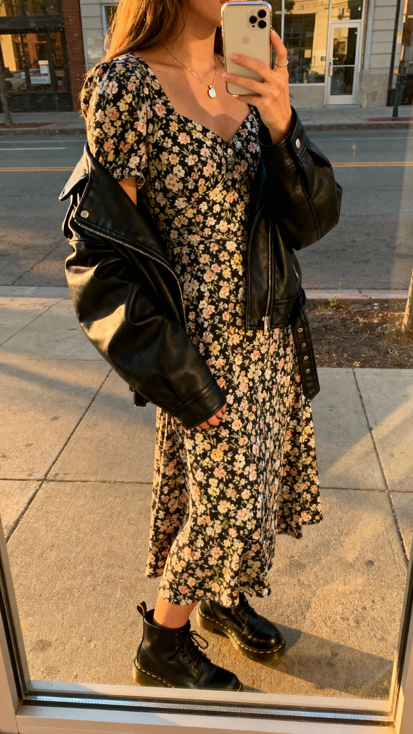 iPhone back-view selfie of a woman wearing a floral midi dress with a black leather jacket draped over shoulders, black Doc Martens, delicate pendant necklace, face not visible, city street window reflection, golden hour daylight, casual iPhone photo.