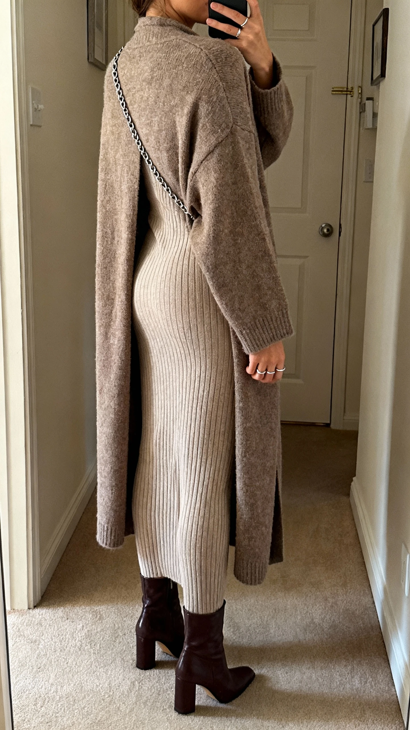 iPhone back-view selfie of a woman wearing a long cardigan coat over a slim knit column dress, heeled boots, crossbody with chain strap, simple rings, face not visible, apartment hallway near door, warm indoor light, natural iPhone photo quality.