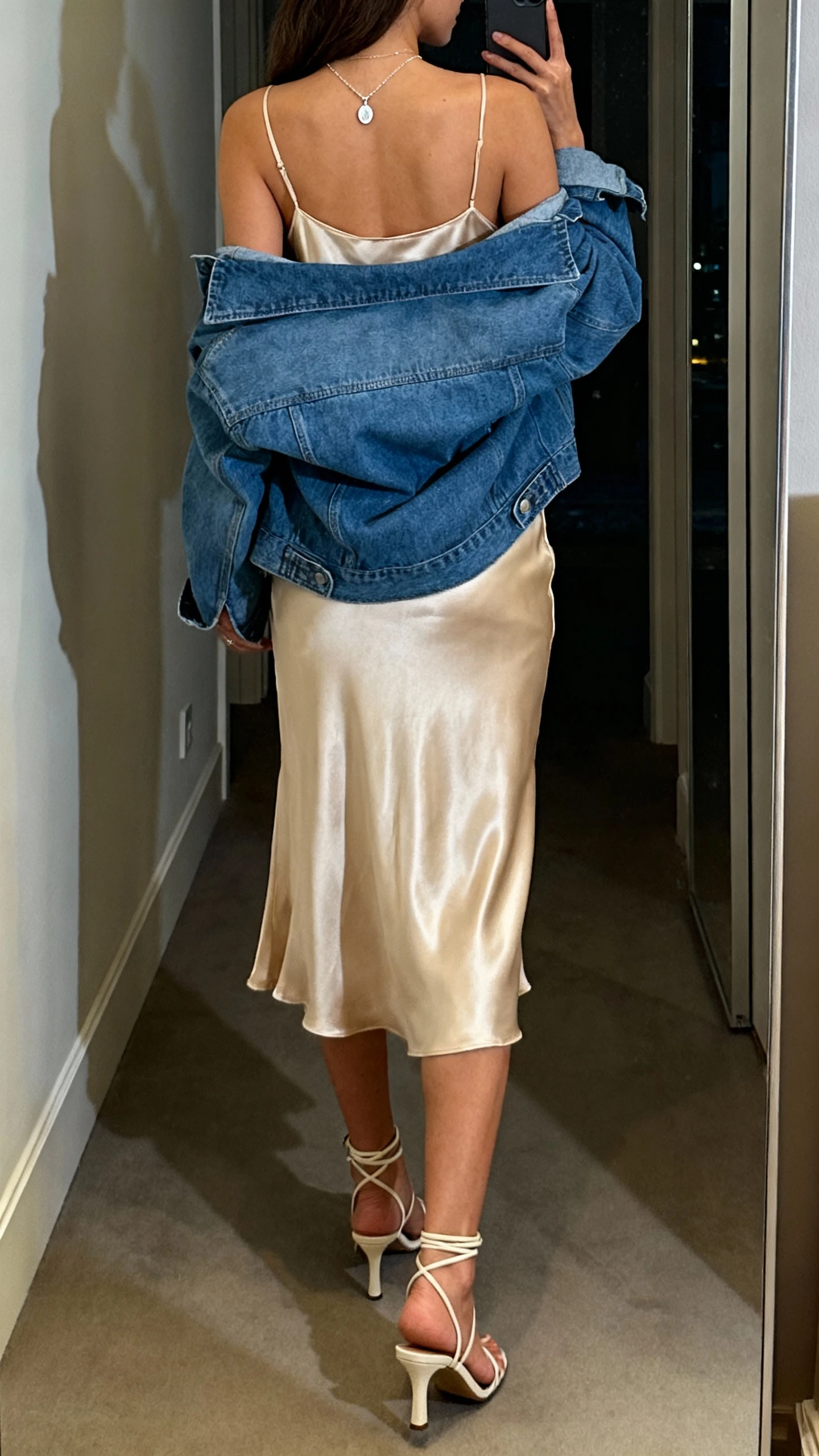 iPhone back-view selfie of a woman wearing a satin slip dress in champagne with an oversized blue denim jacket draped over shoulders, delicate pendant necklace, and strappy heeled sandals; hallway mirror, evening indoor light, face not visible, natural iPhone photo quality.