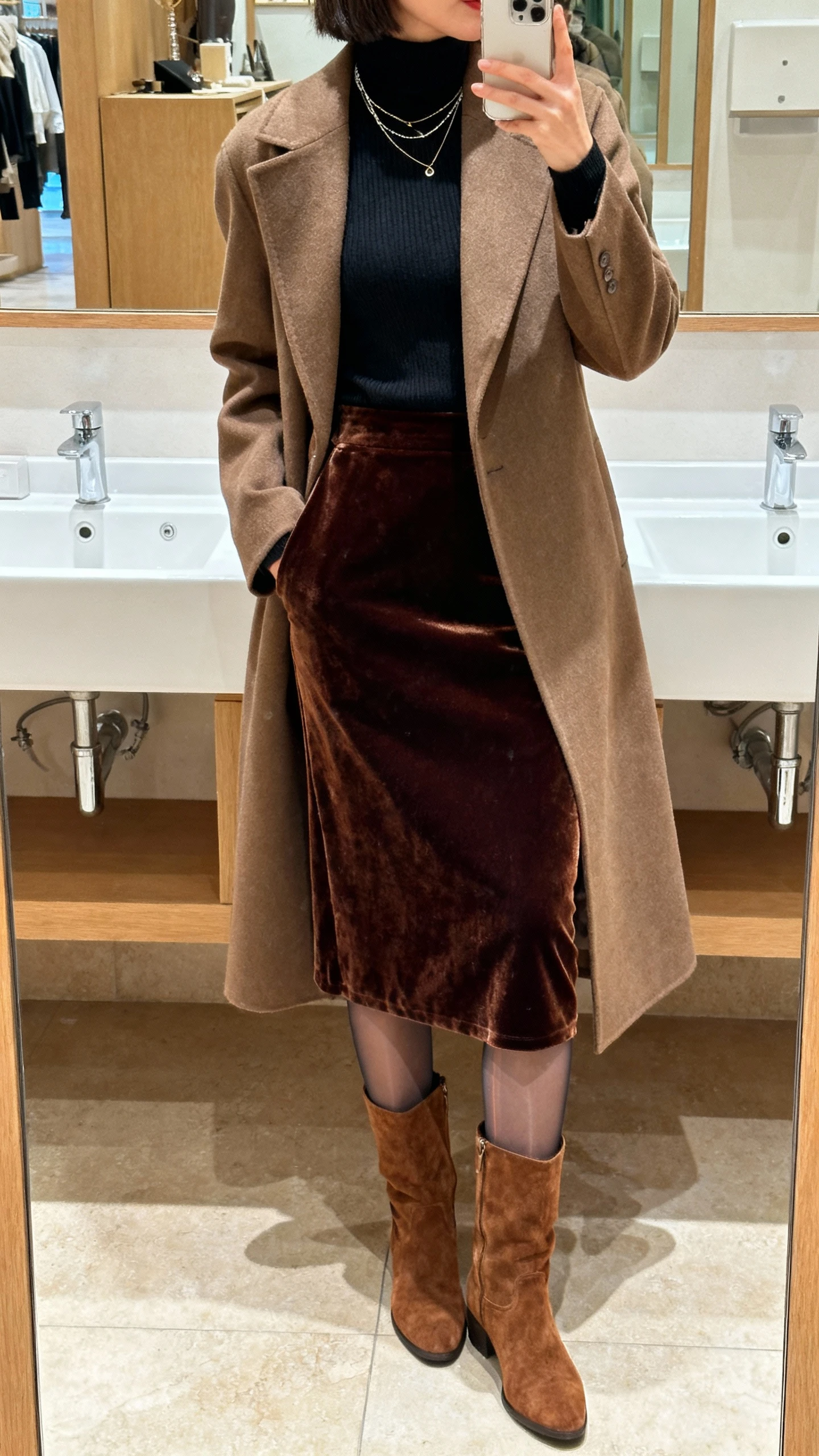 iPhone back-view selfie of a woman wearing a velvet midi skirt, fitted turtleneck, knee-high suede boots, tailored longline coat, sheer thermal tights, delicate layered necklaces peeking at the collar; boutique restroom mirror, face not visible, warm indoor lighting, natural iPhone quality.