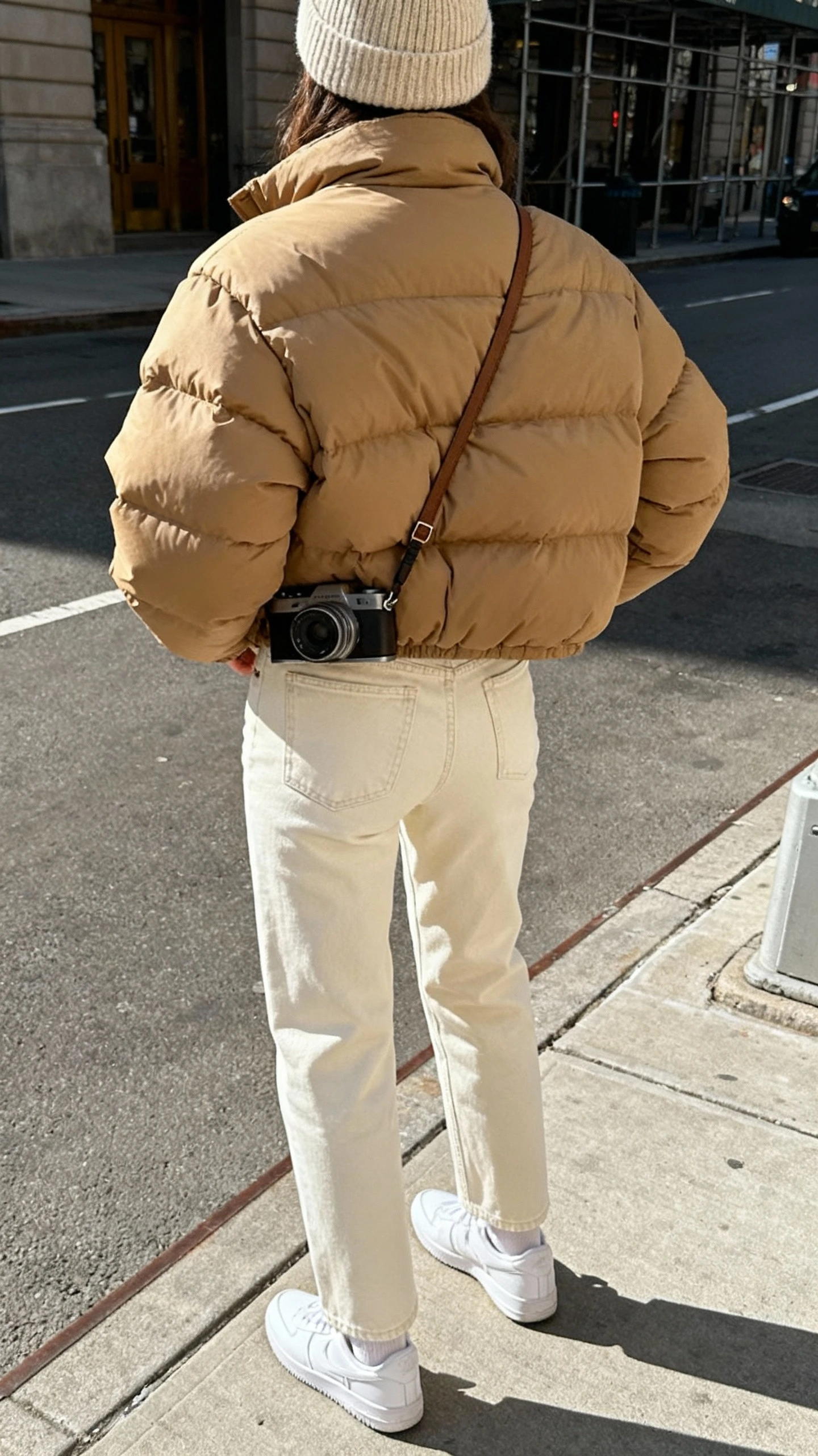 iPhone back-view street selfie of a woman wearing a tan puffer jacket, cream straight-leg denim, white sneakers, knit beanie, crossbody camera bag, face not visible, city sidewalk in daylight, casual iPhone photo.