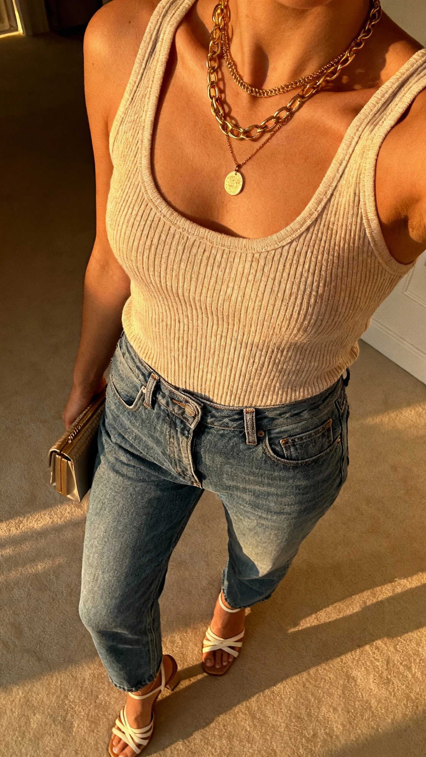 iPhone bar-top selfie cropped at shoulders showing a woman in a fine-knit tank tucked into high-waisted blue jeans, strappy heels, layered gold necklaces, small clutch, face not visible, warm evening indoor lighting, natural iPhone photo.