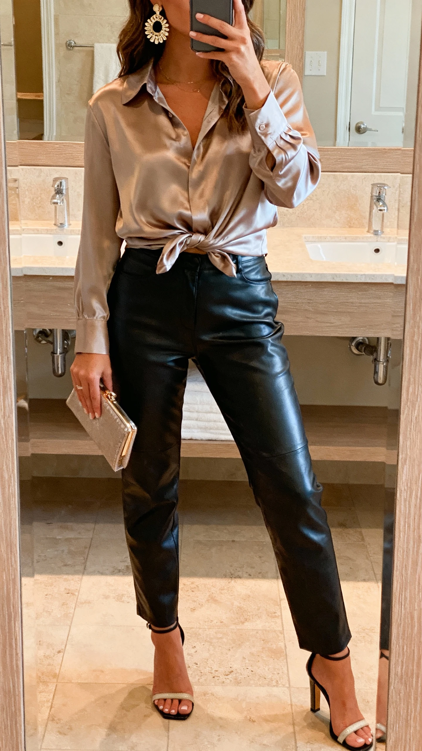 iPhone bathroom mirror selfie of a woman in a satin blouse tucked into leather pants with statement earrings and heeled sandals, small clutch, face not visible, warm vanity lighting, natural iPhone aesthetic.