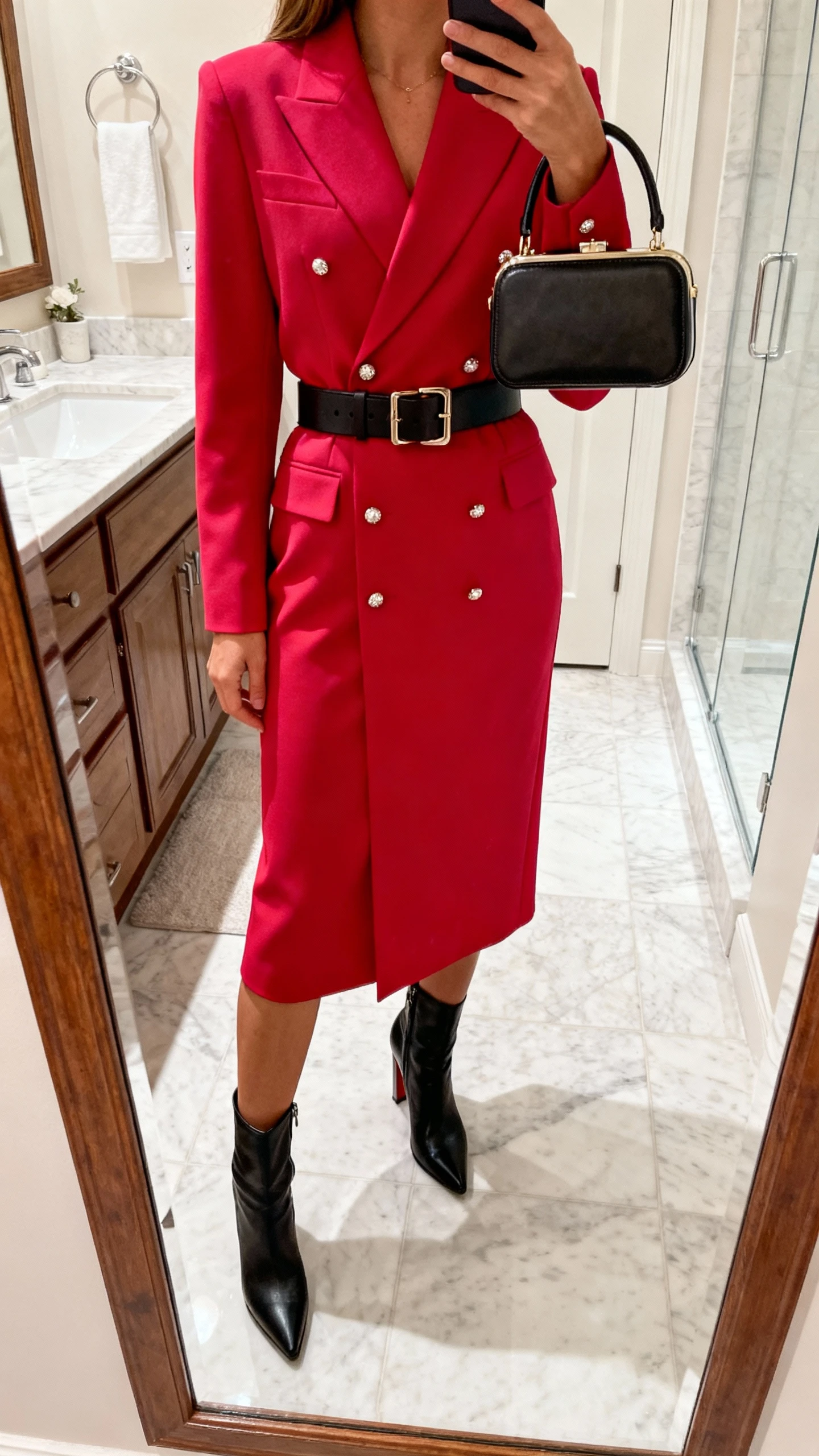 iPhone bathroom mirror selfie of a woman in a statement dress that does the work: bold-colored tailored midi dress, heeled ankle boots, structured belt, simple studs, compact top-handle bag; face not visible, cropped at shoulders, bright indoor lighting, natural iPhone photo quality.