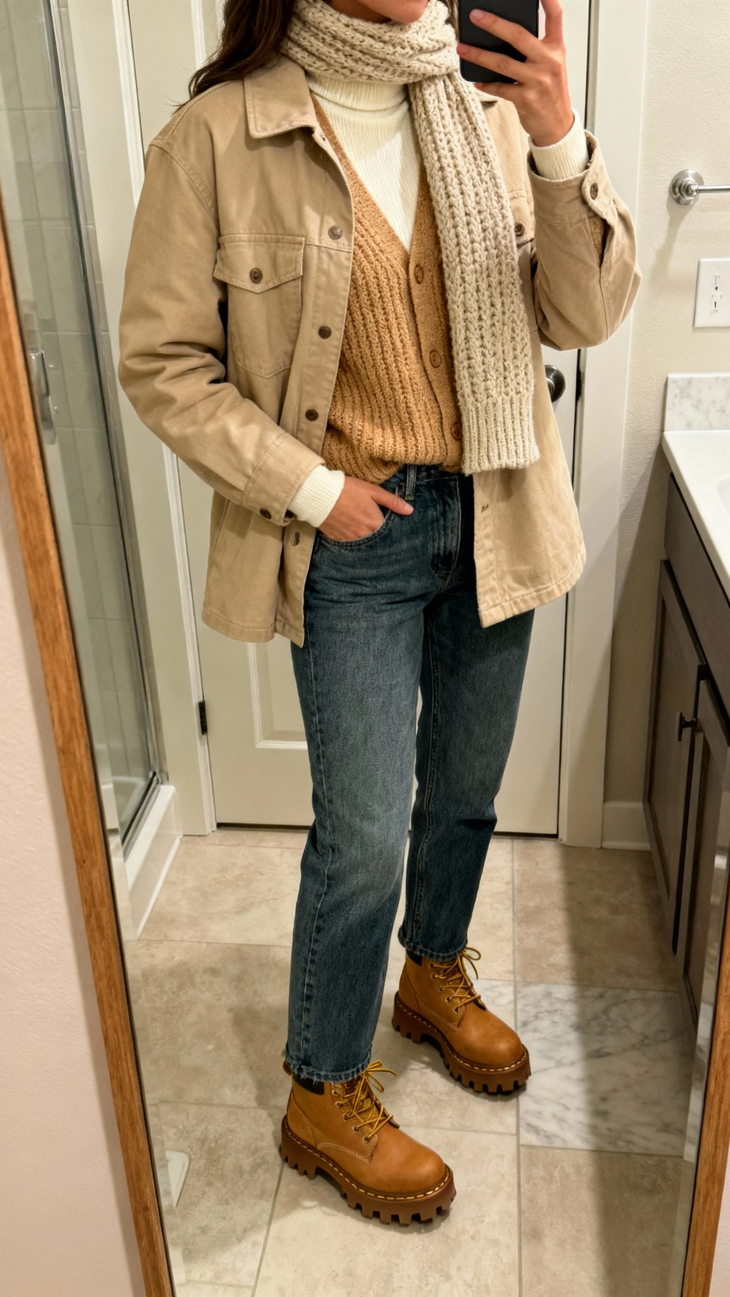 iPhone bathroom mirror selfie of a woman wearing a beige shacket over layered knits (cream turtleneck with a tan chunky cardigan), straight-leg jeans, lug-sole boots, knit scarf, face not visible, cropped at shoulders, warm indoor lighting, natural iPhone photo.