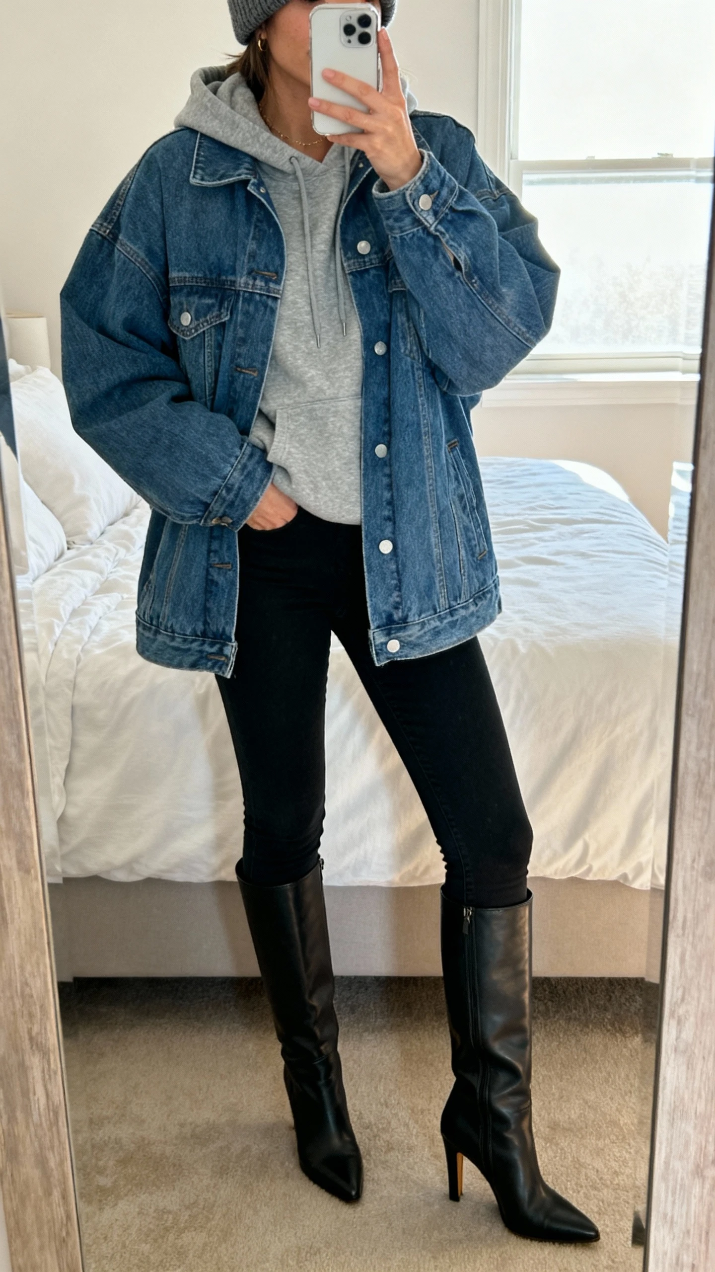 iPhone bedroom mirror selfie of a woman layered in a gray hoodie under an oversized blue denim jacket, black skinny jeans, and knee-high heeled boots to elongate the legs, with a beanie; cropped at neck, soft morning window light, face not visible, natural iPhone photo quality.