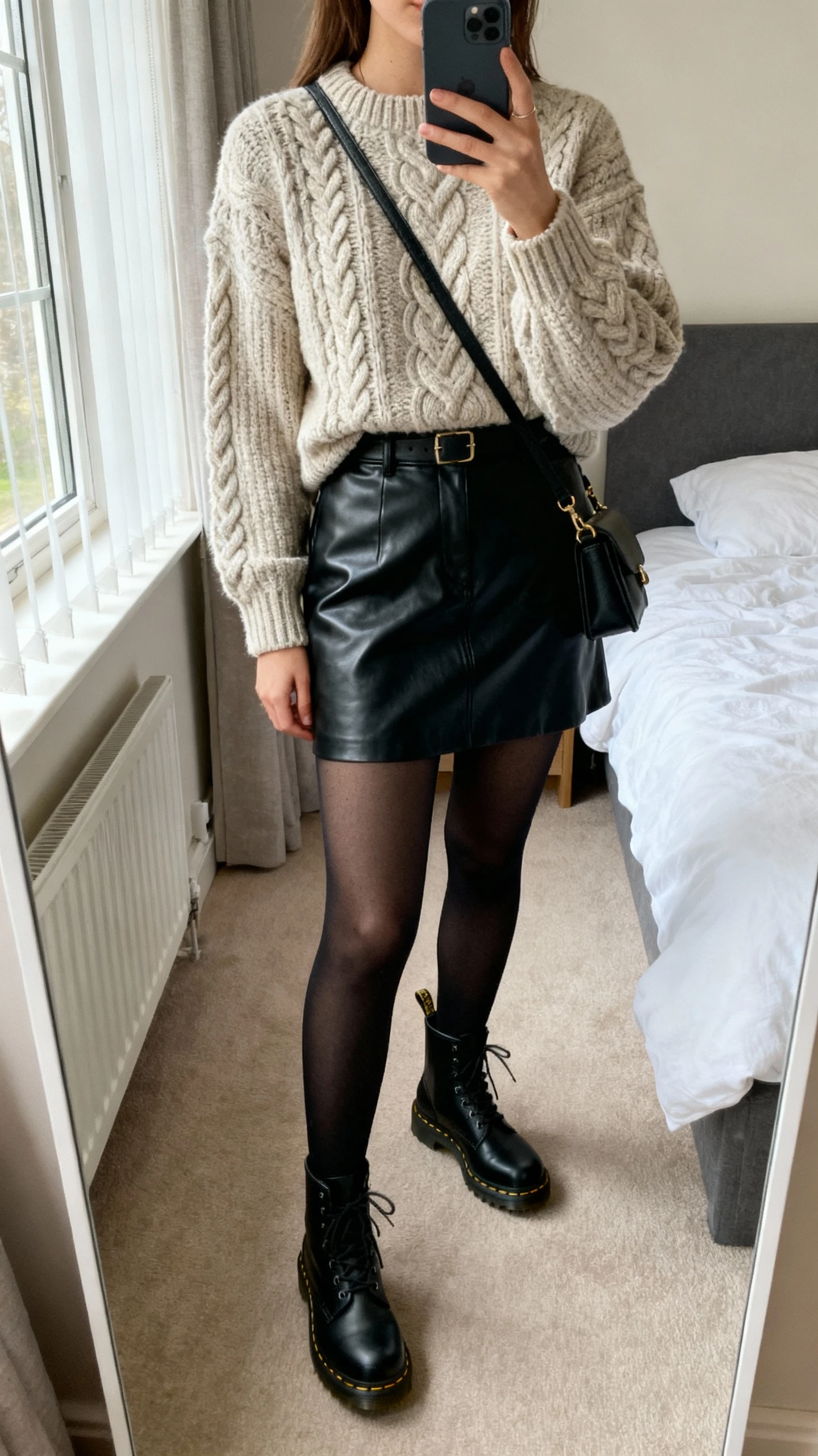iPhone bedroom mirror selfie of a woman wearing a cable-knit sweater tucked into a black leather mini skirt with opaque black tights, black Doc Martens, and a small crossbody, face not visible, cropped at neck, morning window light, natural iPhone photo quality.