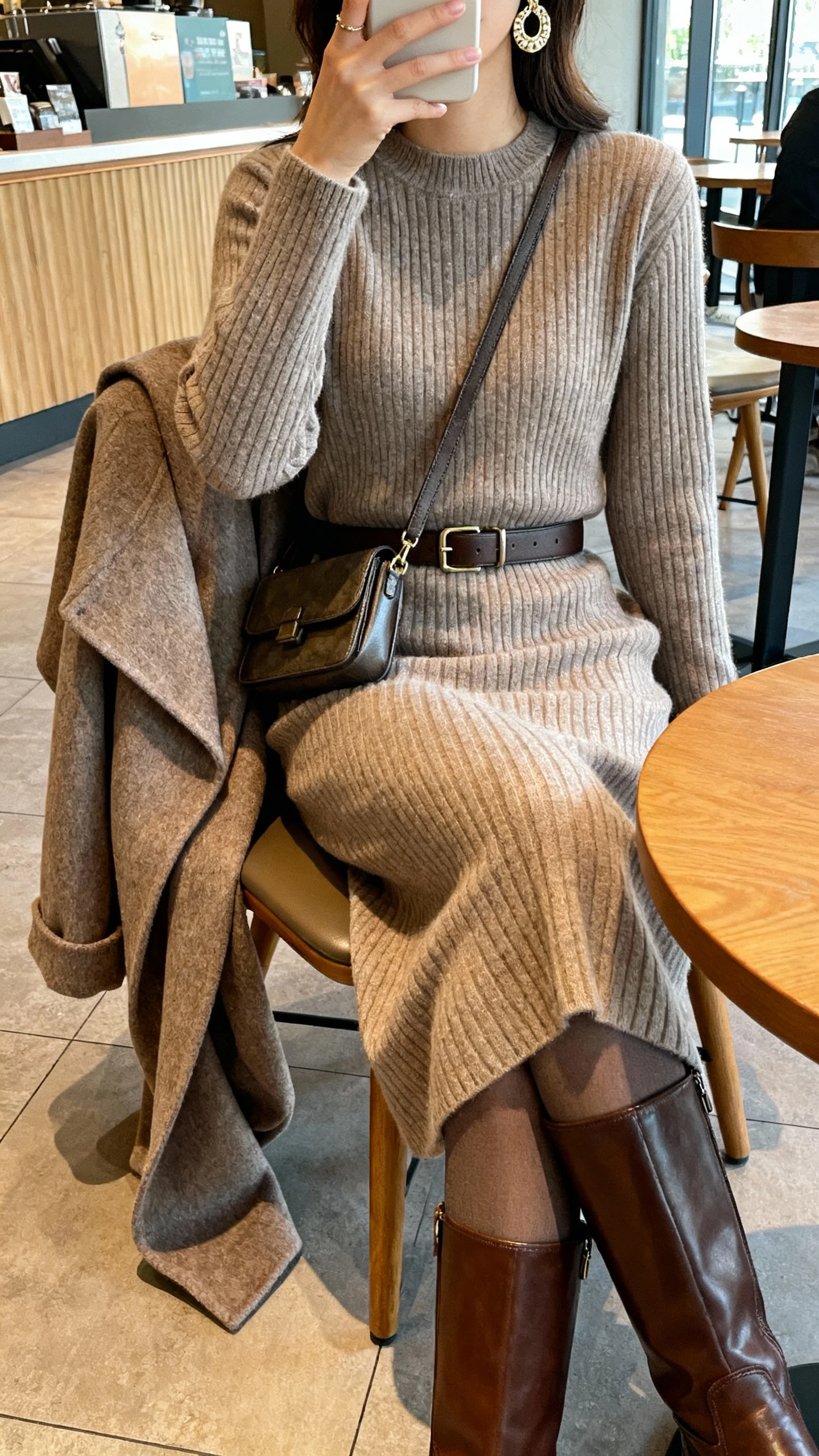 iPhone cafe seated selfie cropped at shoulders showing a woman in a ribbed midi sweater dress with a medium-width belt, leather knee-high boots, a wool wrap coat draped over the chair, opaque tights, a structured crossbody, and statement earrings; face not visible, warm indoor light, natural iPhone photo quality.