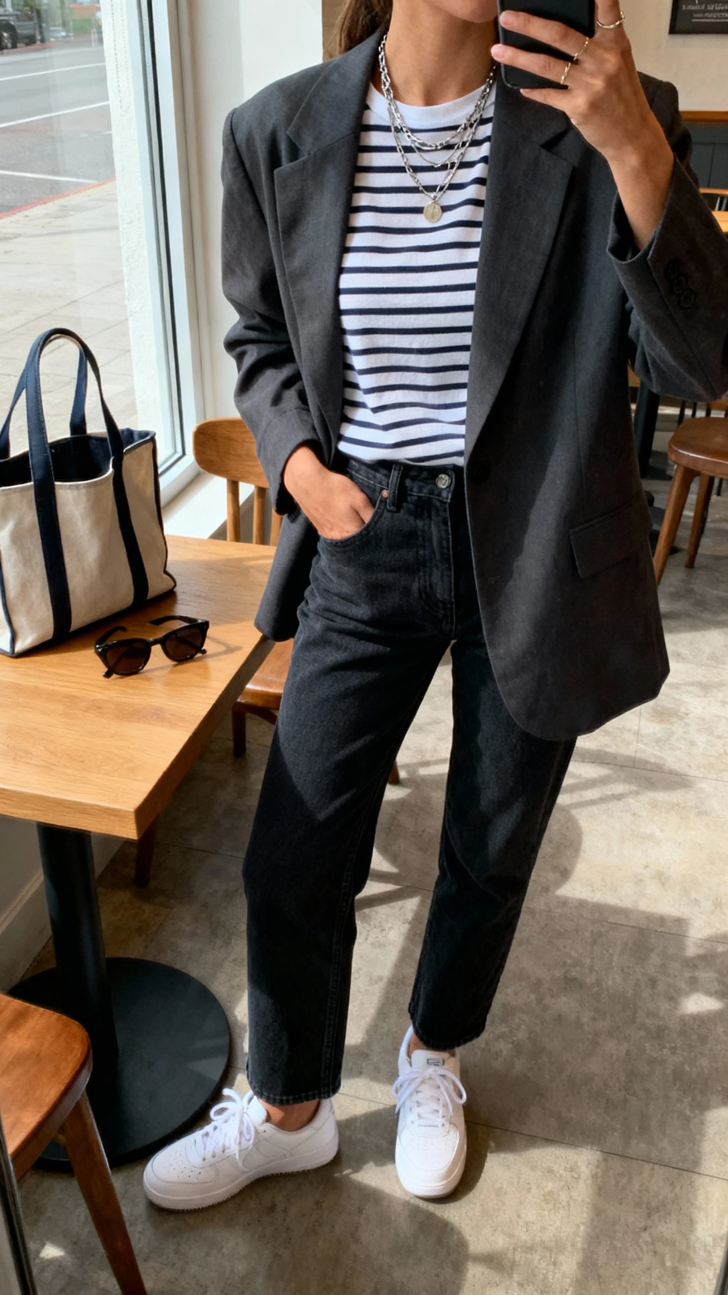 iPhone cafe table selfie cropped at neck of a woman in smart-casual Friday uniform: relaxed blazer, striped tee, dark straight-leg jeans, white sneakers, layered necklaces, tote and sunglasses on table; face not visible, window light, natural iPhone photo quality.