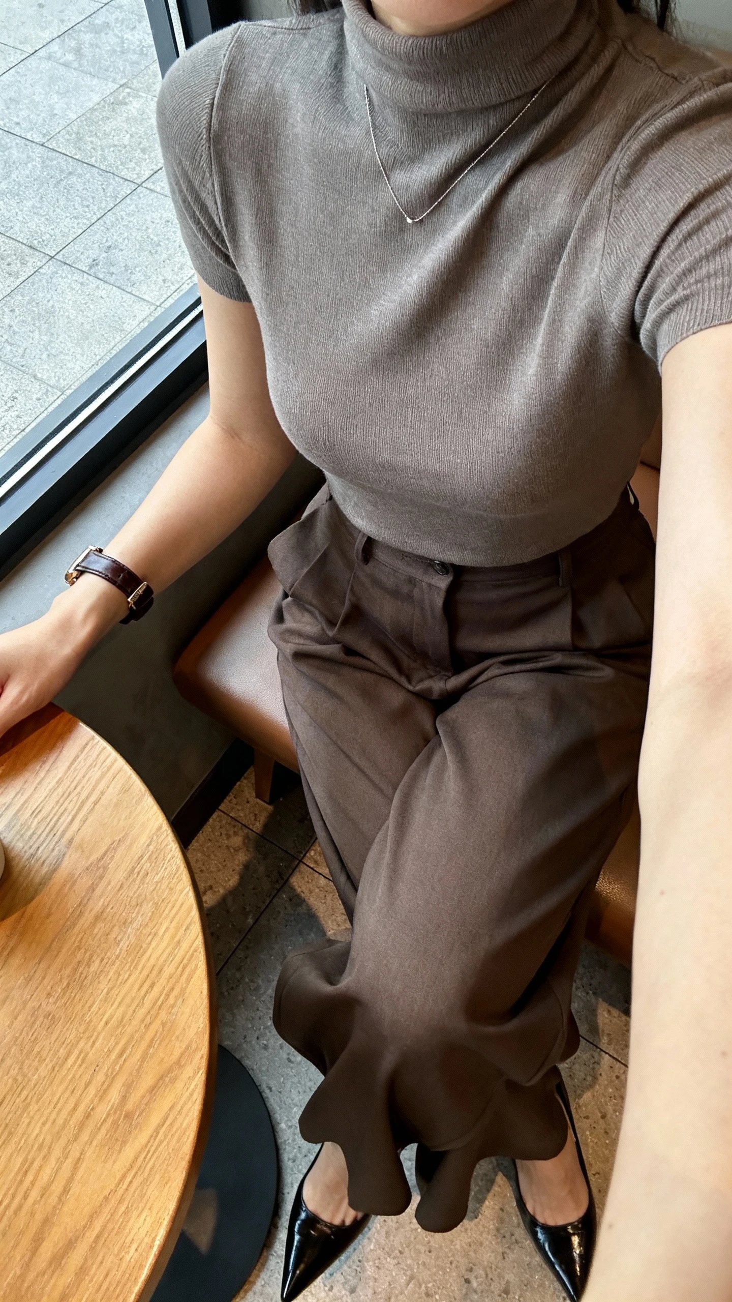 iPhone cafe table selfie cropped at shoulders of a woman wearing a sleek turtleneck tucked into high-waisted wide-leg trousers, pointed heels, minimalist necklace, leather watch, face not visible, window-side cafe seat, natural daylight, natural iPhone photo quality.