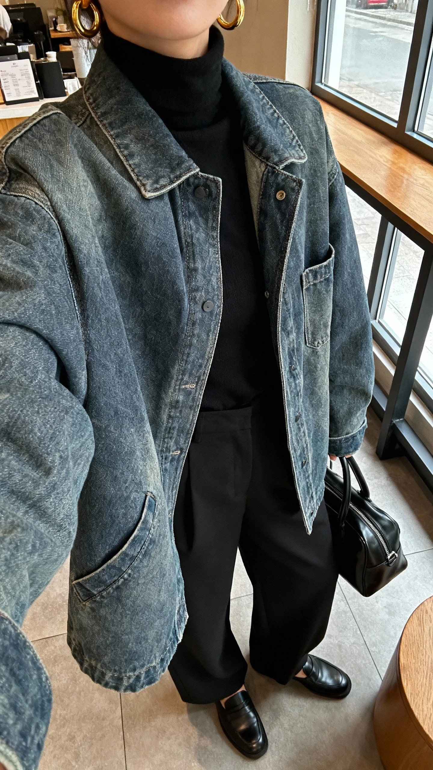 iPhone cafe table selfie cropped at shoulders showing a woman in an all-black outfit—black turtleneck, black wide-leg trousers, black loafers—and a medium-wash oversized denim jacket for contrast, with gold hoops and a black leather tote; face not visible, window light in coffee shop, natural iPhone photo quality.