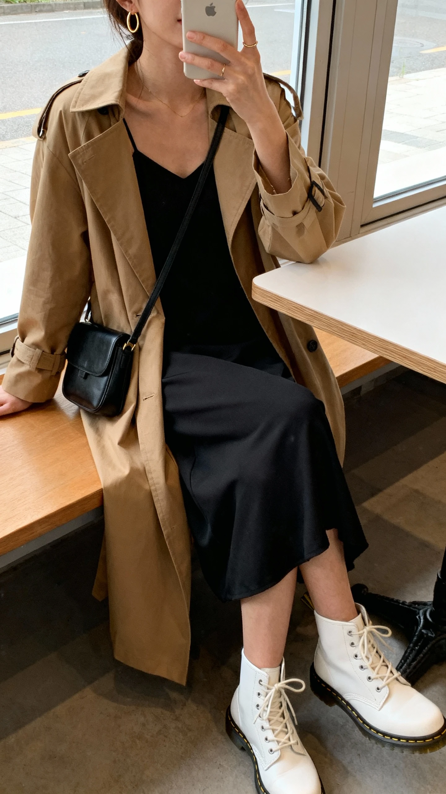 iPhone cafe table selfie, side angle, of a woman in a minimalist dress and trench: black midi slip dress, tan trench coat, white Doc Martens, small black shoulder bag, thin gold hoops; face not visible, window seat with soft daylight, natural iPhone photo quality.