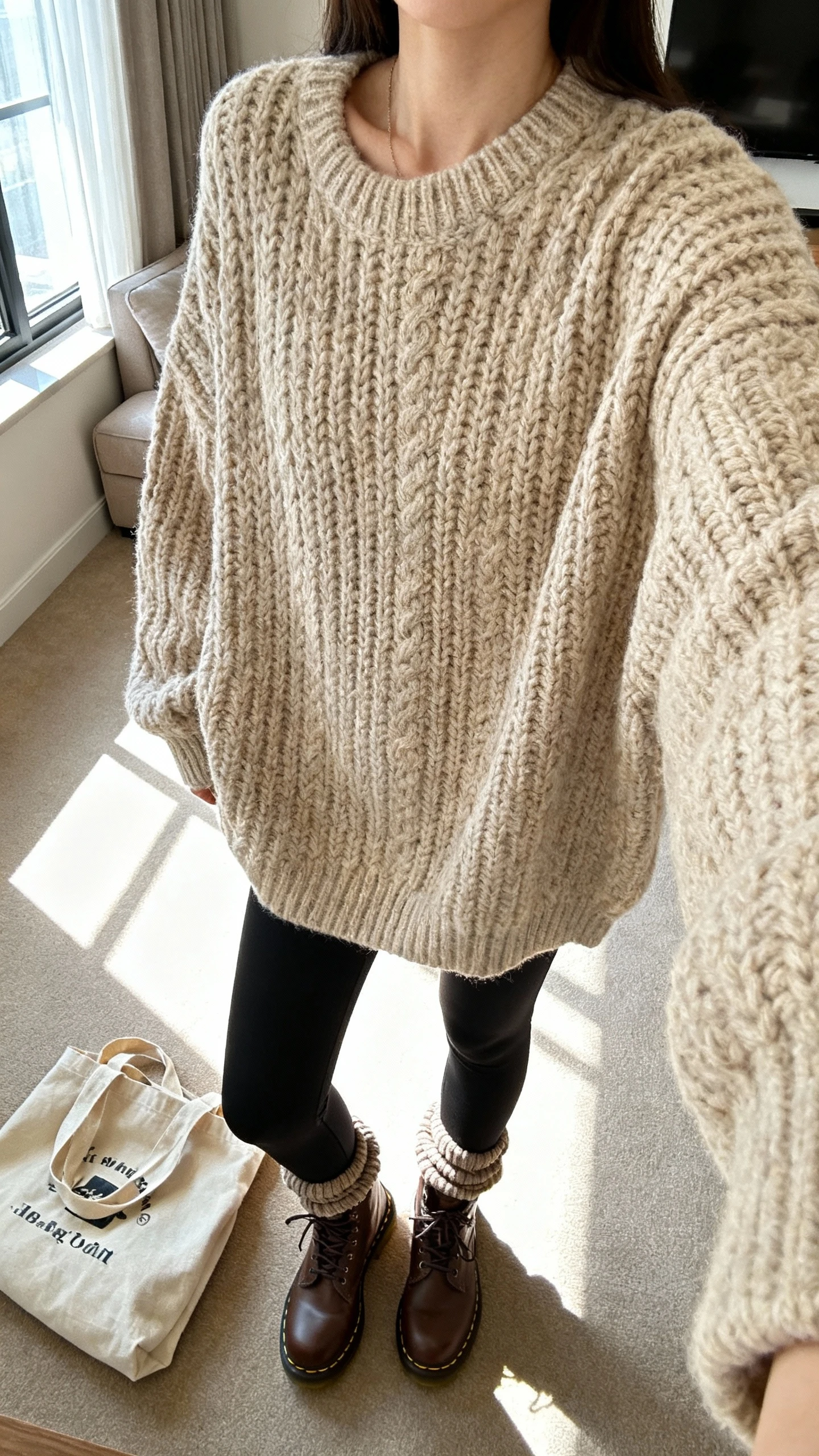 iPhone couch selfie cropped at neck showing a woman in an oversized chunky beige knit, thick black leggings, brown Doc Martens, scrunched socks, and a tote bag on the floor; face not visible, living room window light, cozy iPhone photo.