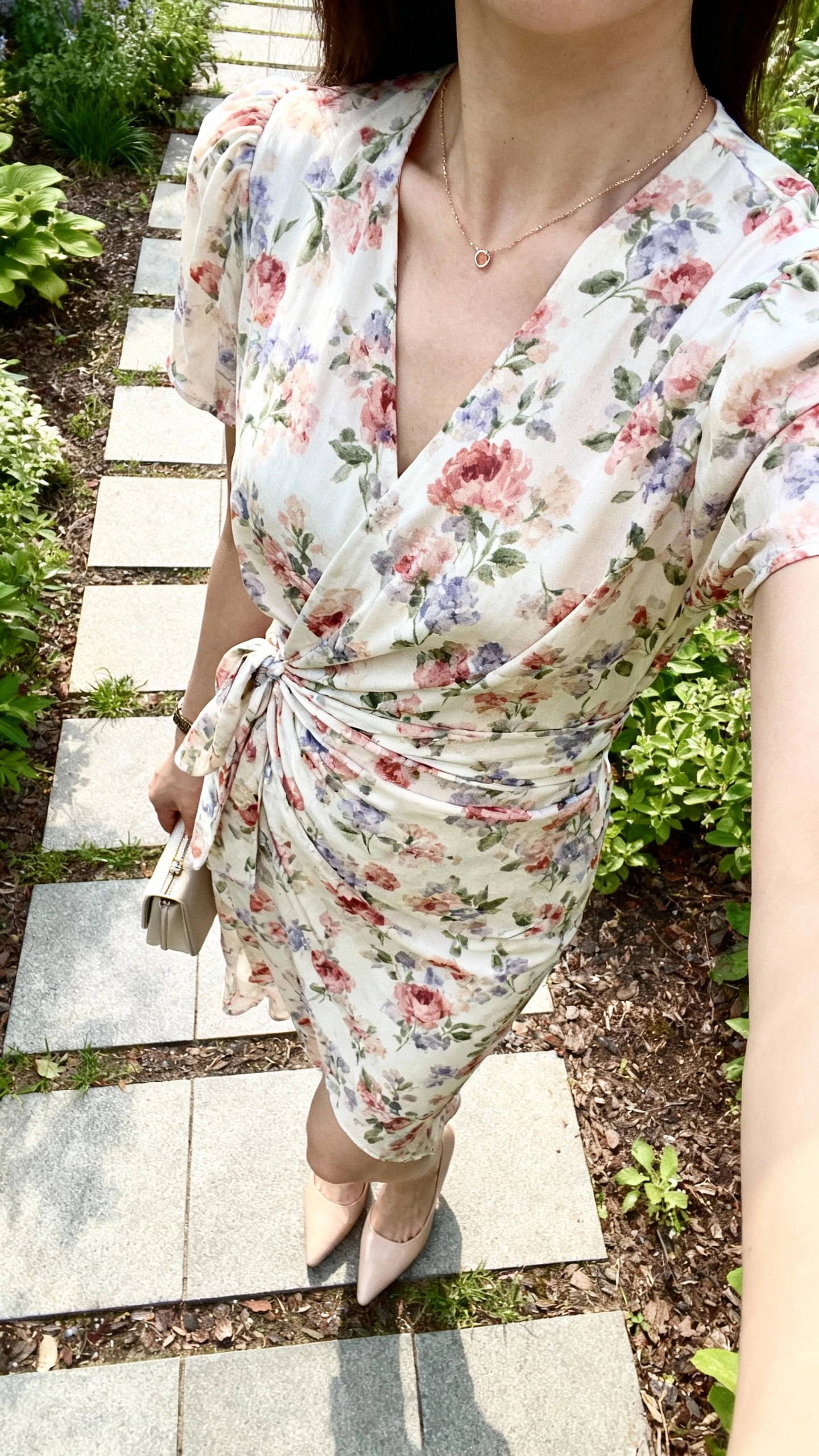 iPhone cropped-at-shoulders selfie of a woman wearing a floral wrap dress with low heels, dainty necklace, small clutch, face not visible, garden path setting in gentle daylight, casual iPhone aesthetic.