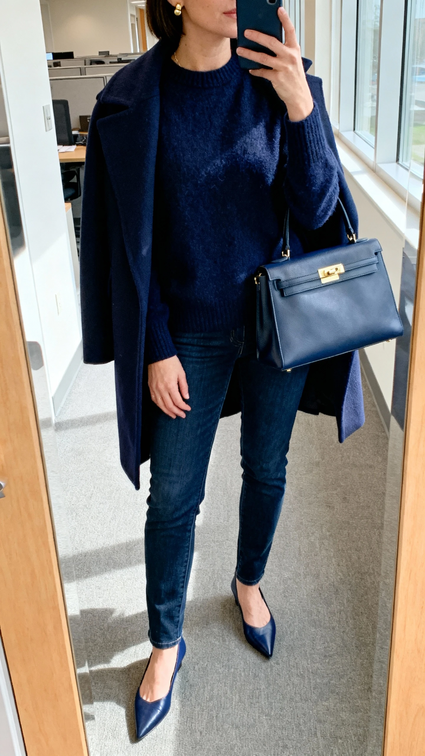 iPhone cropped-neck selfie showing a woman in a monochrome navy look: navy cashmere sweater, dark blue jeans, navy wool coat draped over shoulders, navy pointed flats, structured navy handbag, minimal gold studs, face not visible, office hallway mirror, bright morning light, iPhone photo.