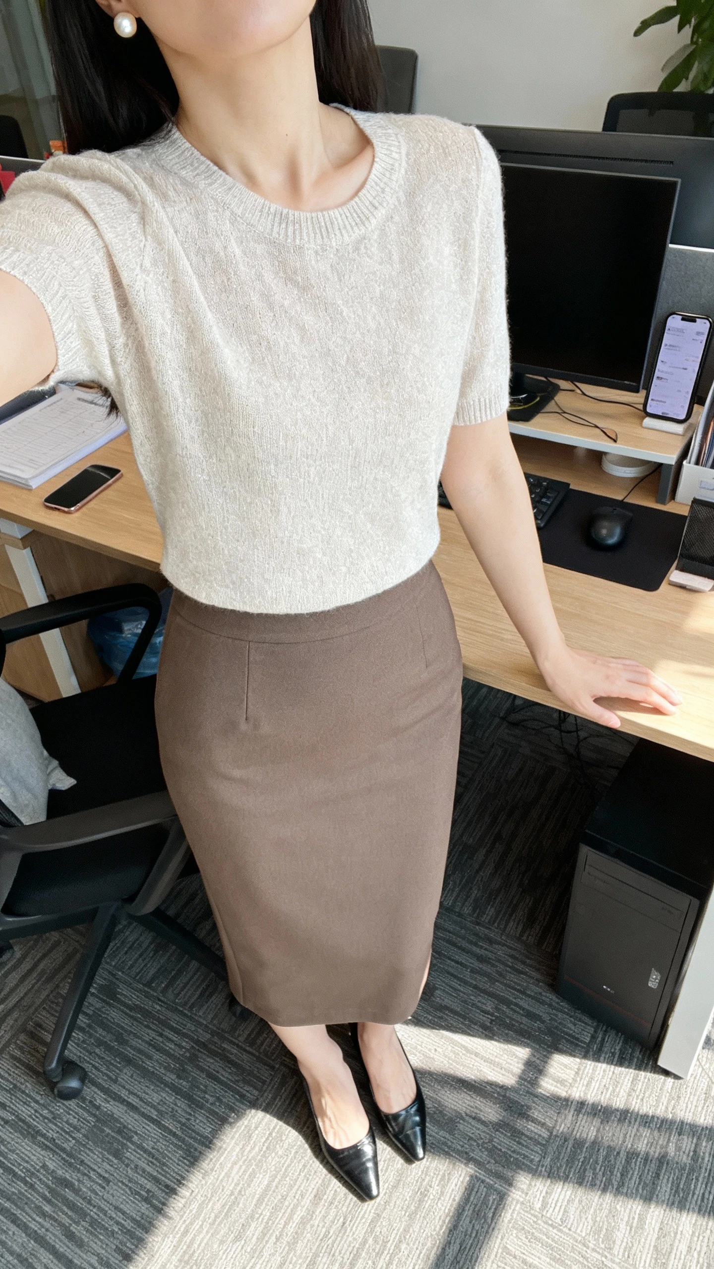 iPhone desk selfie cropped at neck showing a woman in a midi pencil skirt and a soft knit top, classic pumps, pearl studs, face not visible, office setting with morning light, natural iPhone photo quality.