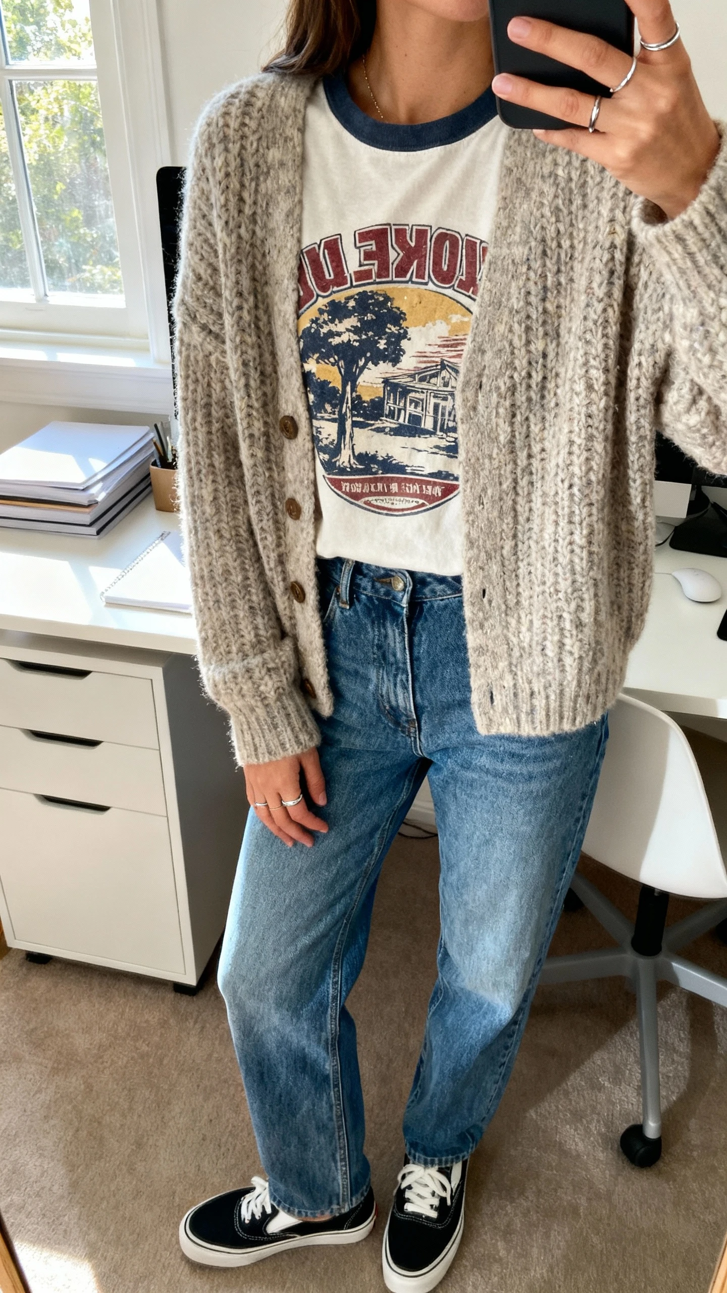 iPhone desk selfie cropped at shoulders showing a woman wearing a vintage-style graphic tee under a chunky cardigan with relaxed blue jeans, slip-on sneakers, simple rings, face not visible, home office setting, soft afternoon window light, natural iPhone photo.