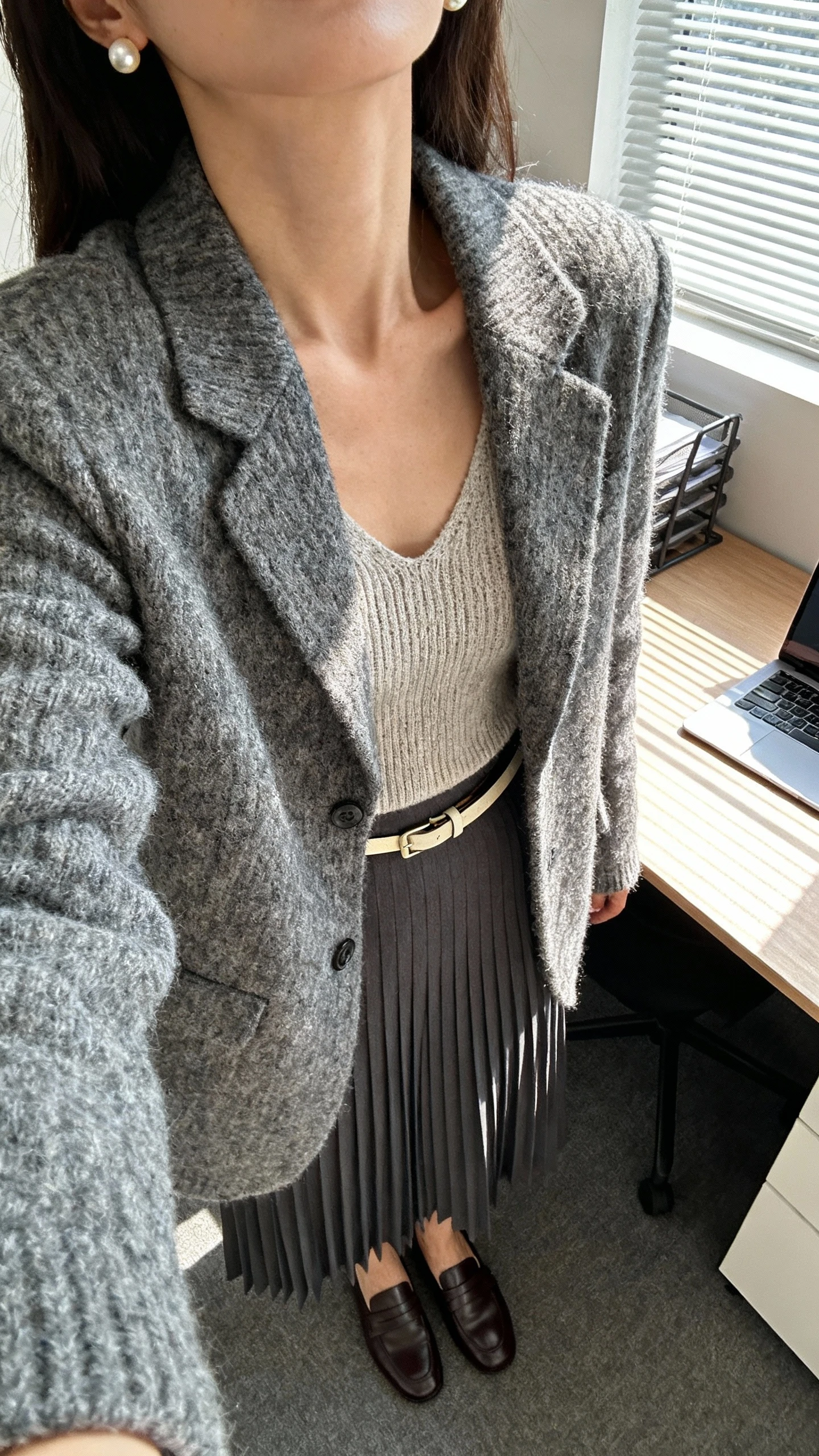 iPhone desk-side selfie cropped at neck showing a woman wearing a sweater blazer over a knit shell with a pleated skirt, loafers, thin belt, pearl studs, face not visible, office setting with window light, natural iPhone photo.