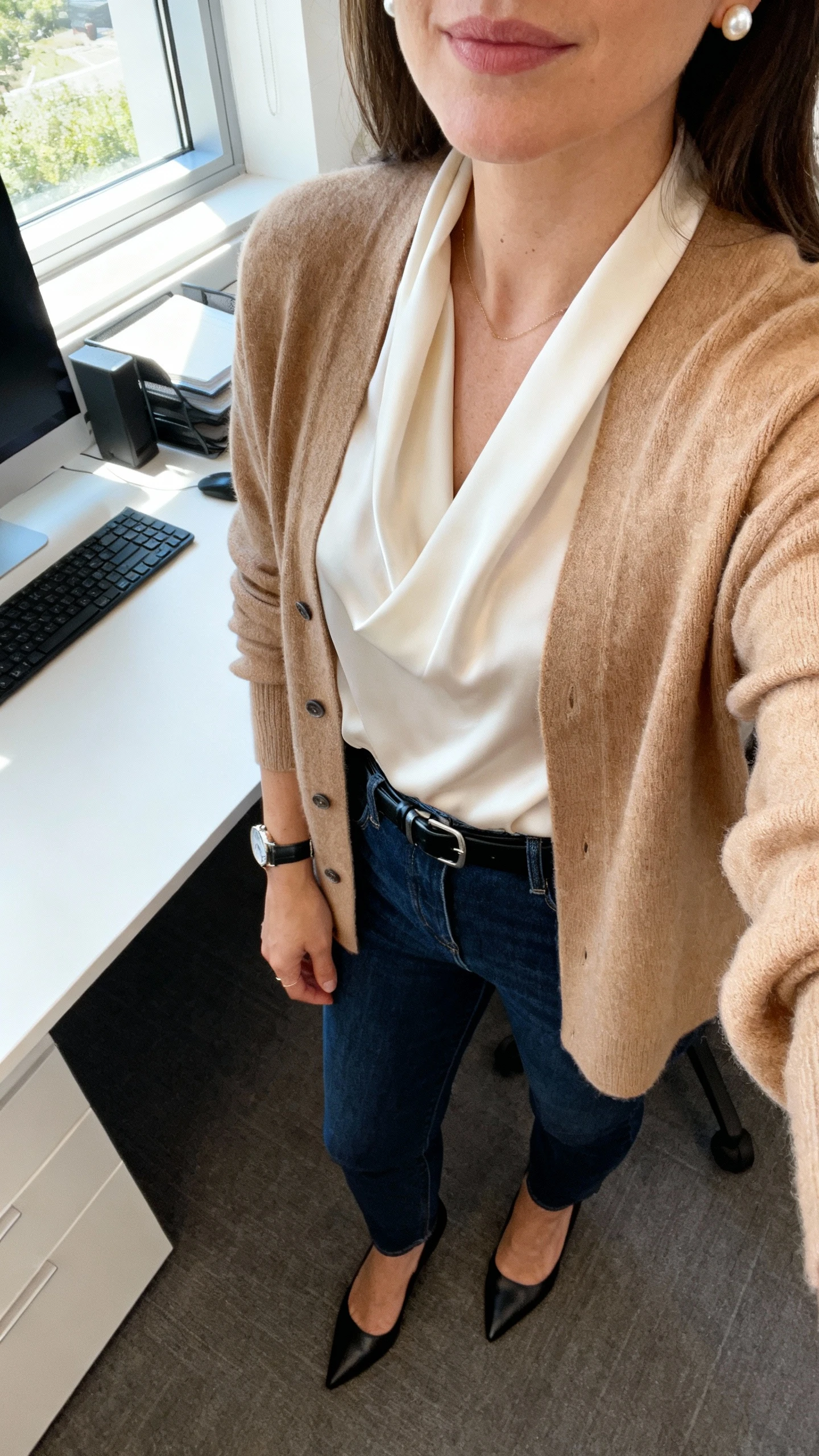 iPhone desk-side selfie cropped at shoulders of a woman wearing a soft camel cardigan over a silk ivory blouse, dark blue jeans, black pumps, slim black belt, pearl studs and a watch, face not visible, modern office setting, morning window light, natural iPhone photo quality.