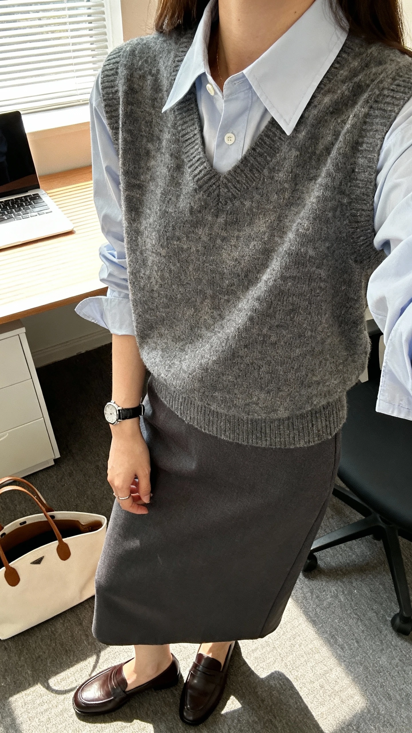 iPhone desk-side selfie cropped at shoulders showing a woman in a sweater vest layered over a crisp button-up shirt with a midi skirt, loafers, watch, tote, face not visible, office setting, morning window light, natural iPhone photo quality.