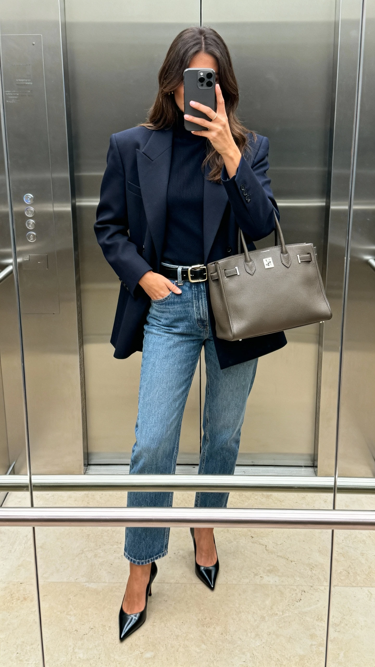 iPhone elevator mirror selfie of a woman in elevated denim with a statement blazer, sleek belt, pointed-toe heels, structured tote, face not visible, phone covering face, indoor lighting, natural iPhone aesthetic.