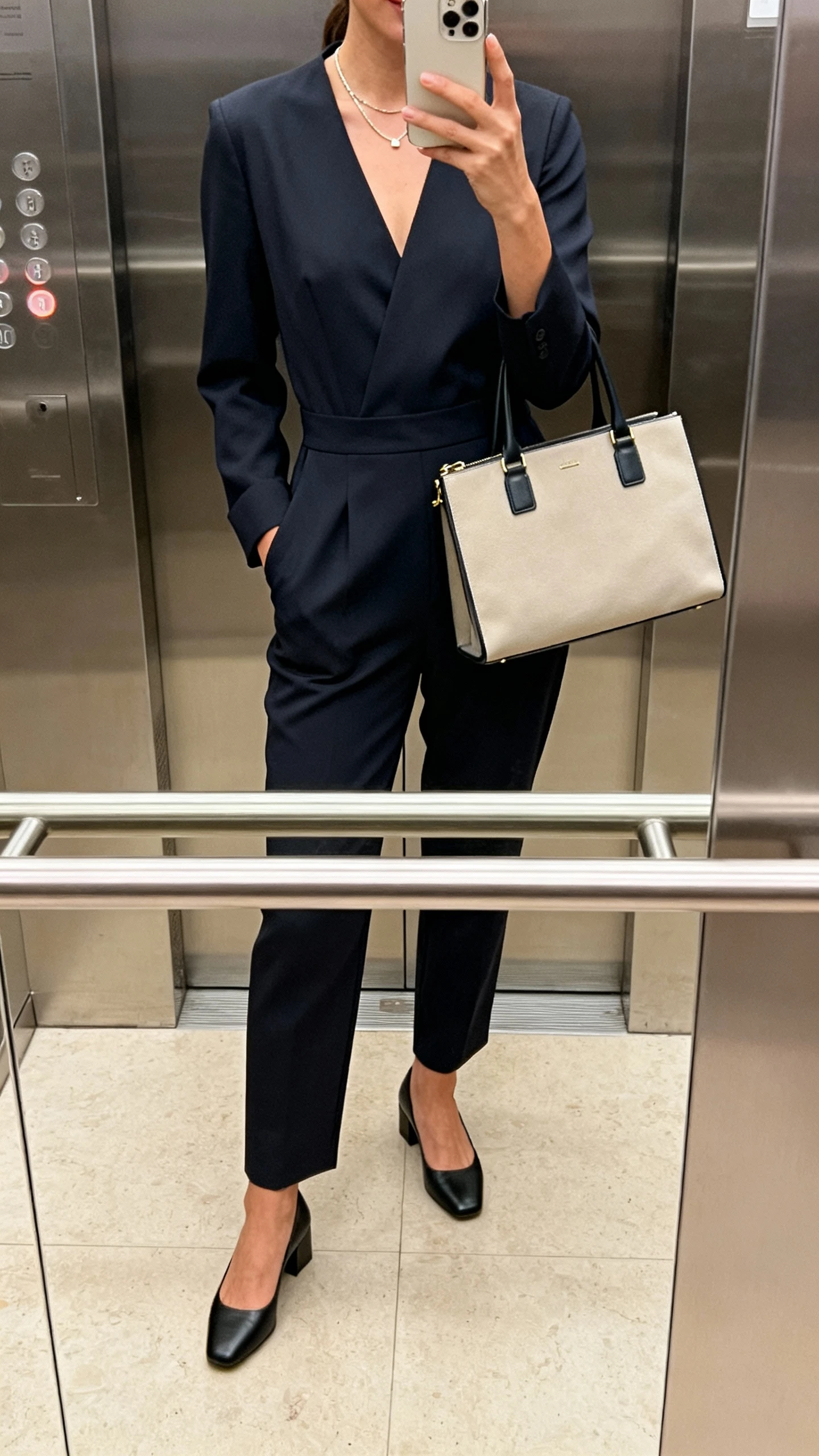 iPhone elevator mirror selfie of a woman wearing a smart tailored jumpsuit, low block heels, minimalist necklace, and a work tote, face not visible, natural indoor lighting, everyday iPhone look