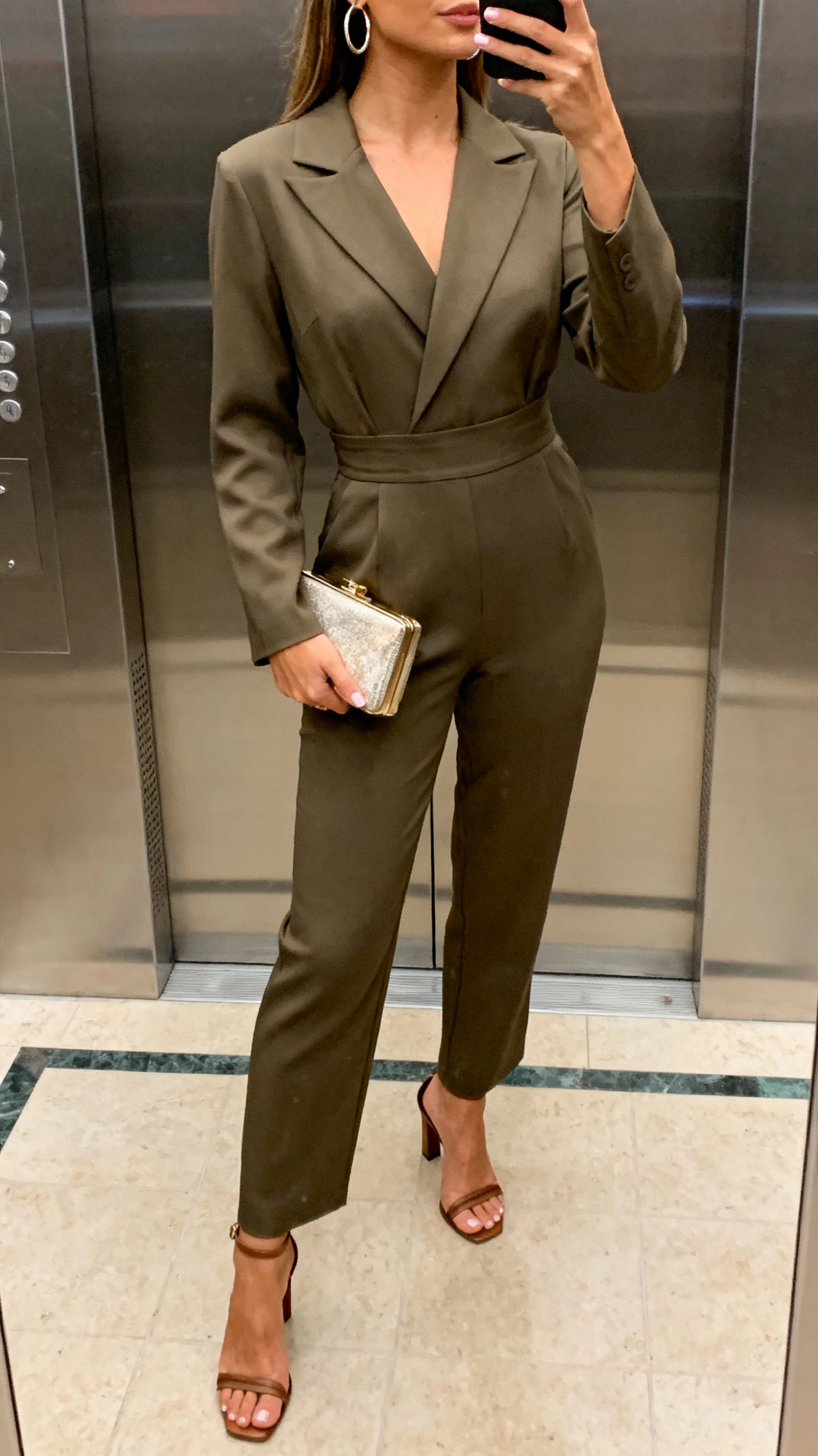 iPhone elevator mirror selfie of a woman wearing a tailored jumpsuit with a defined waist, heeled sandals, hoop earrings, and a compact clutch, face not visible, cropped at neck, ambient indoor lighting, natural iPhone quality.