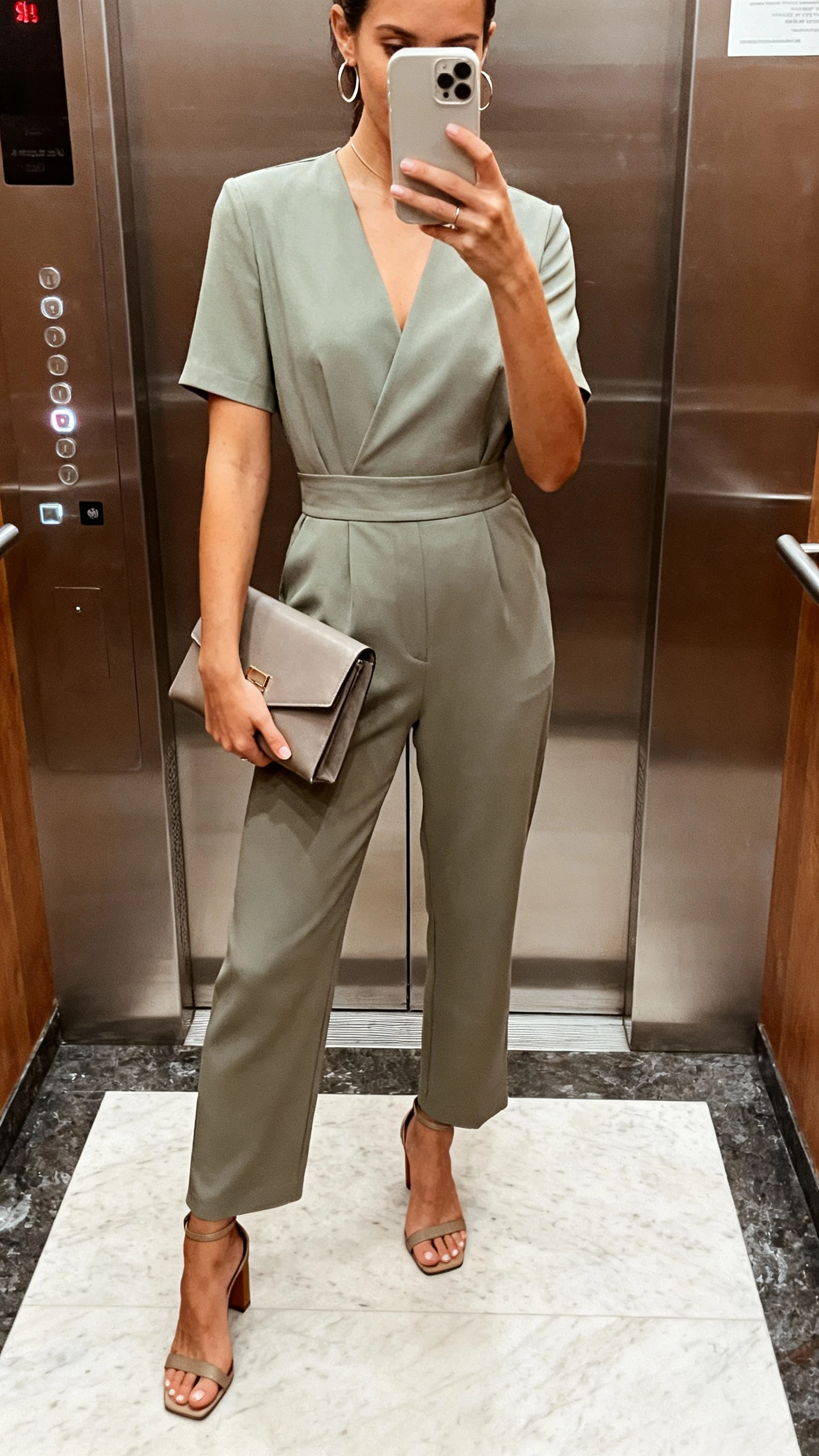 iPhone elevator selfie of a woman wearing a minimal tailored jumpsuit with a defined waist, heeled sandals, structured clutch, thin hoop earrings, face not visible, indoor lighting, natural iPhone photo quality.