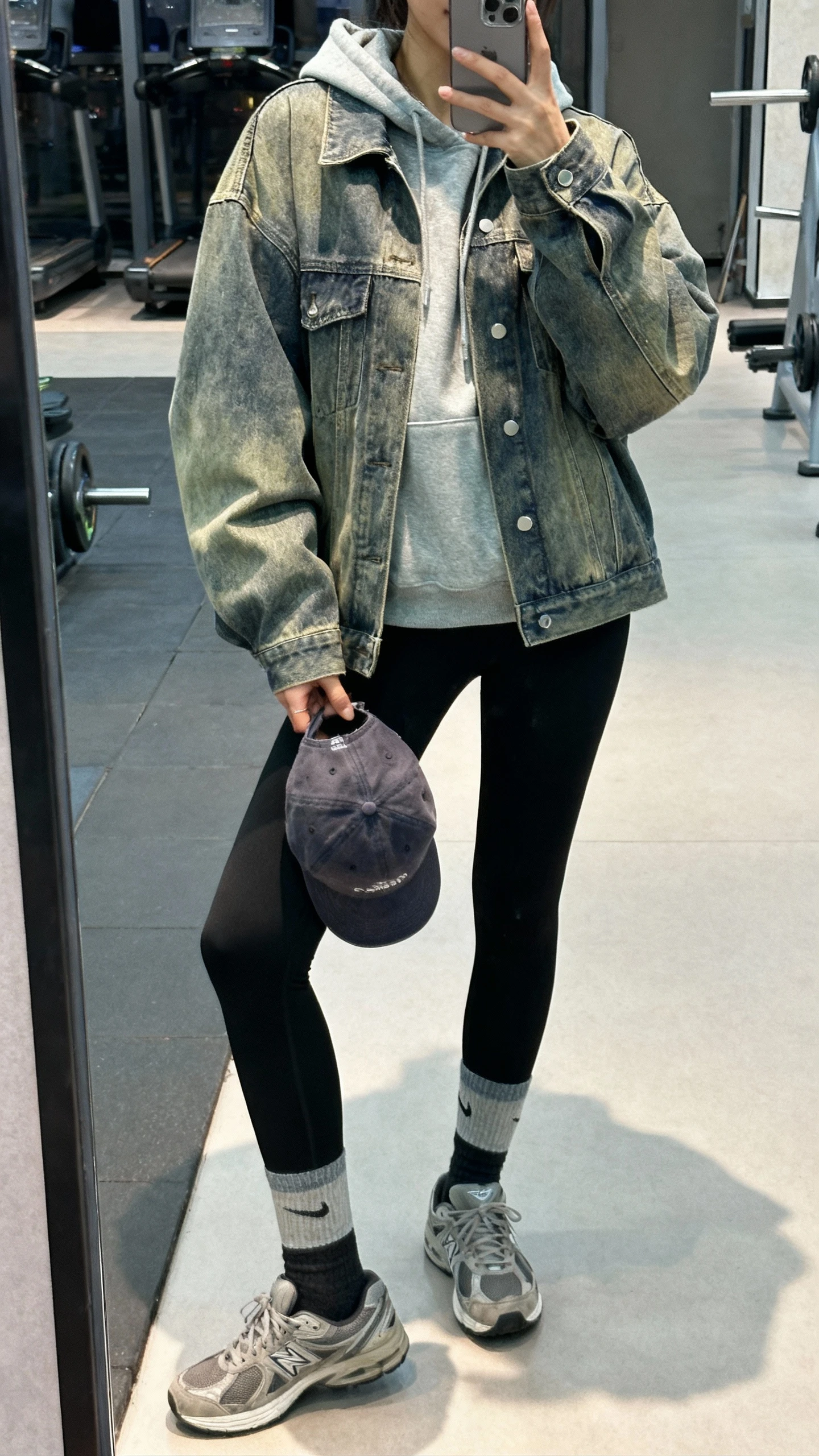 iPhone gym-mirror selfie of a woman wearing black leggings, a gray hoodie, and a boxy mid-wash denim jacket, sporty socks and sneakers, baseball cap in hand, face not visible, natural indoor lighting, casual iPhone aesthetic