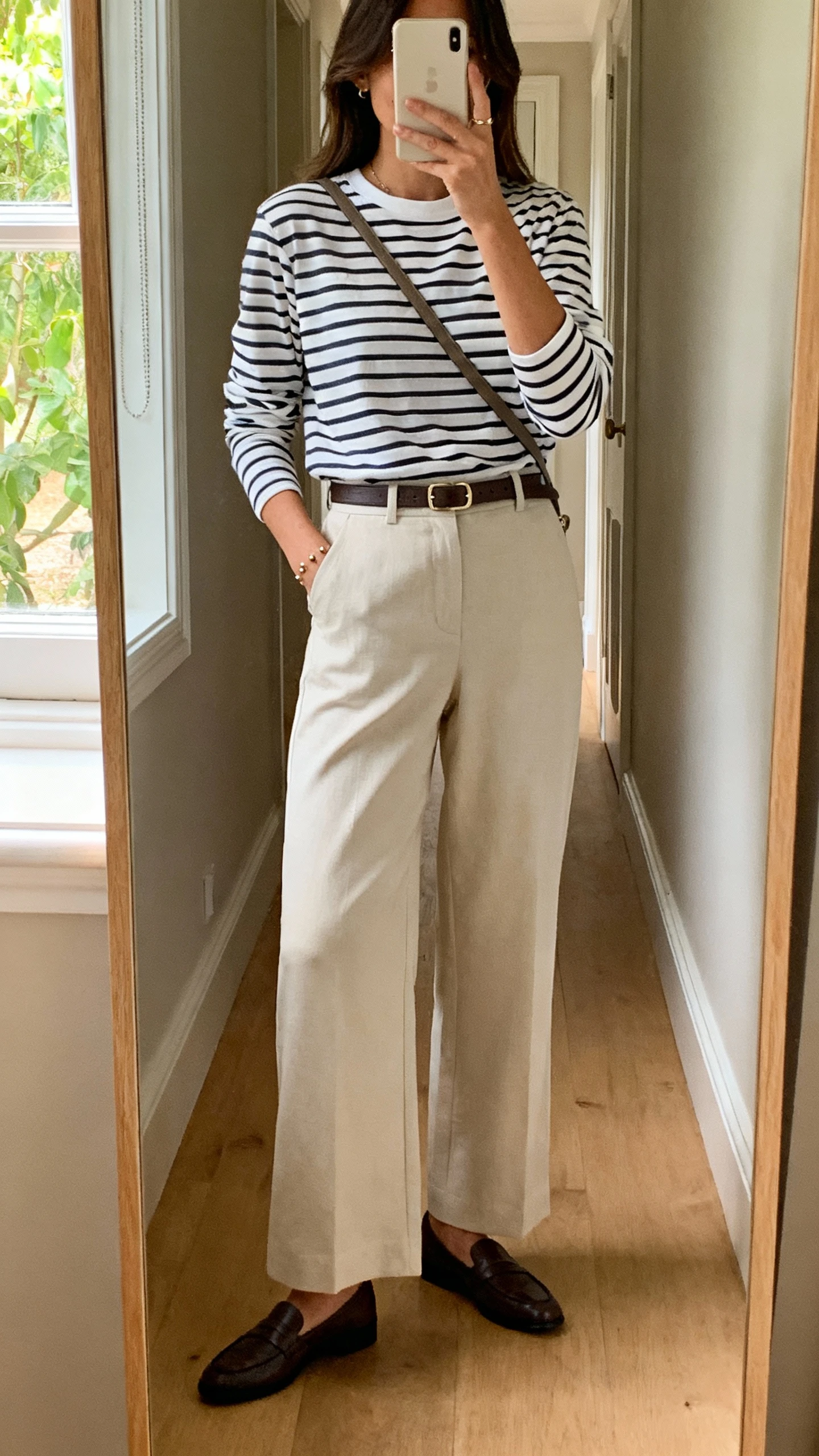 iPhone hallway mirror selfie of a woman in a Breton striped long-sleeve tee tucked into high-waisted wide-leg trousers, loafers, a slim belt, simple studs, and a crossbody, face not visible, natural daylight from window, casual iPhone aesthetic.