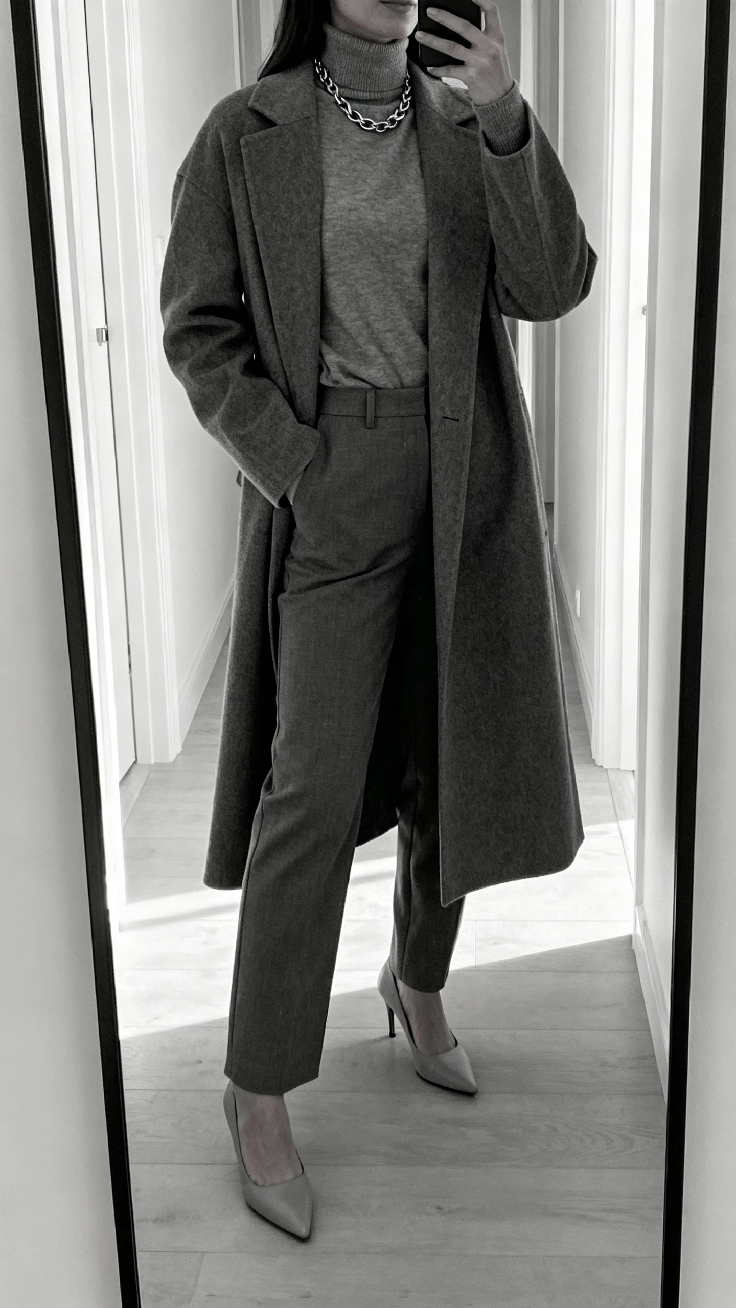 iPhone hallway mirror selfie of a woman in a monochrome magic set: same-tone turtleneck and tailored trousers, matching longline coat, tonal pumps, subtle chain necklace; face not visible, full-length mirror, morning daylight, natural iPhone photo quality.