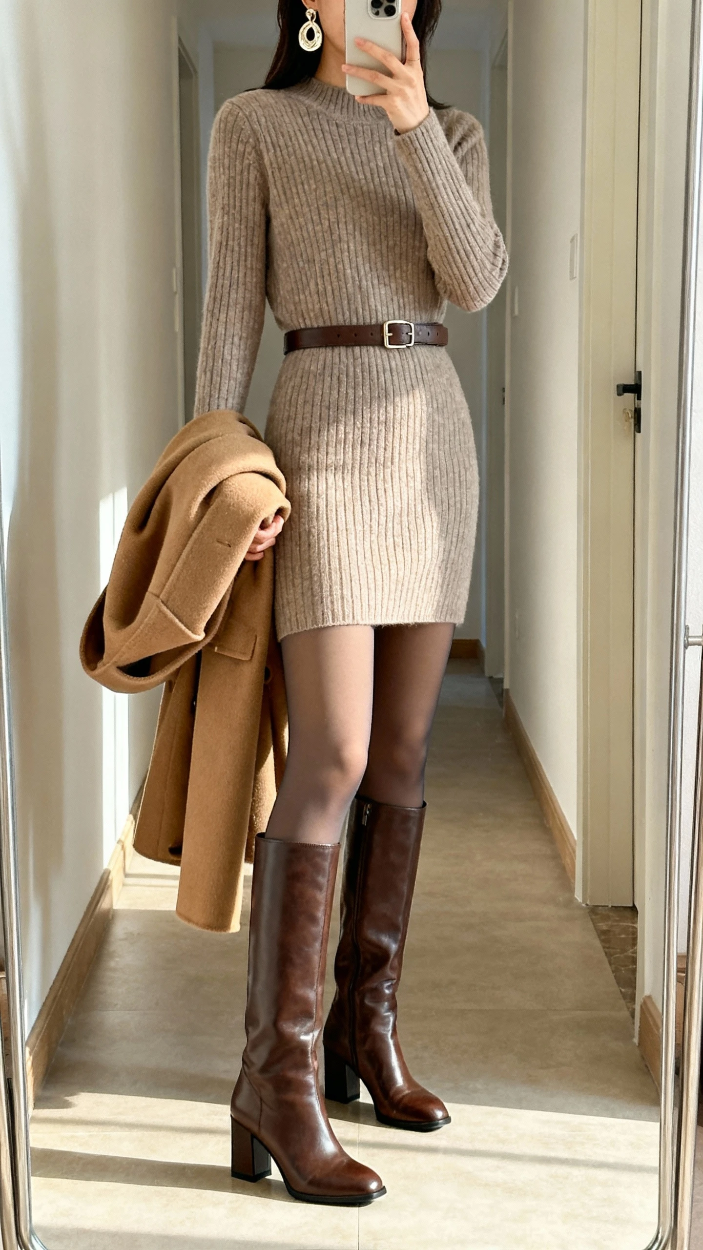 iPhone hallway mirror selfie of a woman wearing a knee-length ribbed cashmere sweater dress, opaque thermal tights (80 denier), knee-high leather boots with block heel, structured camel wool coat draped over arm, leather belt at waist, statement earrings; face not visible, natural indoor light, casual iPhone aesthetic.