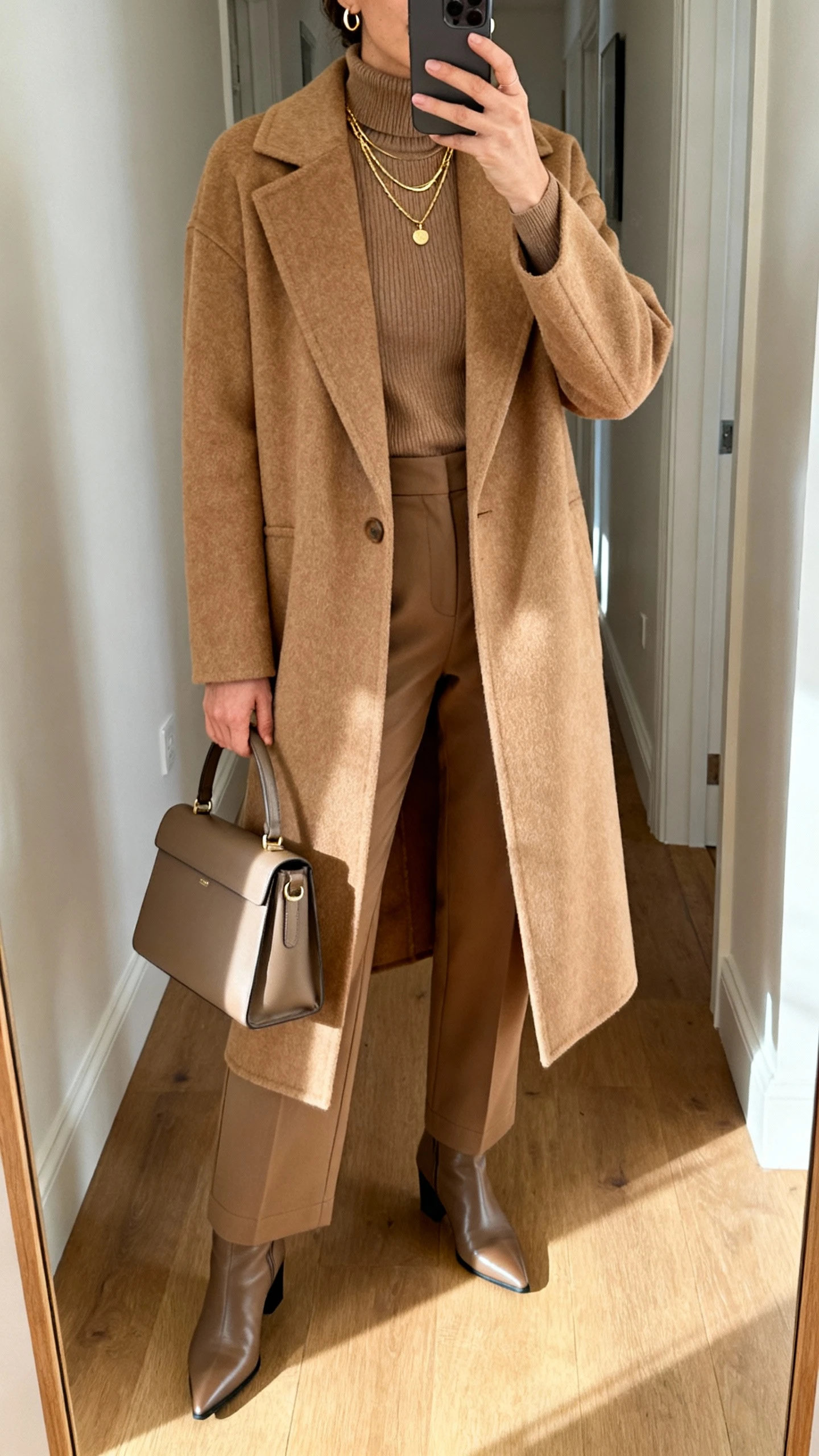 iPhone hallway mirror selfie of a woman wearing a long camel wool-blend coat over a fitted merino turtleneck, high-waisted straight-leg trousers, pointed ankle boots, a structured top-handle bag, delicate gold layered necklaces and small hoops; cropped at neck, morning light, face not visible, natural iPhone photo quality.