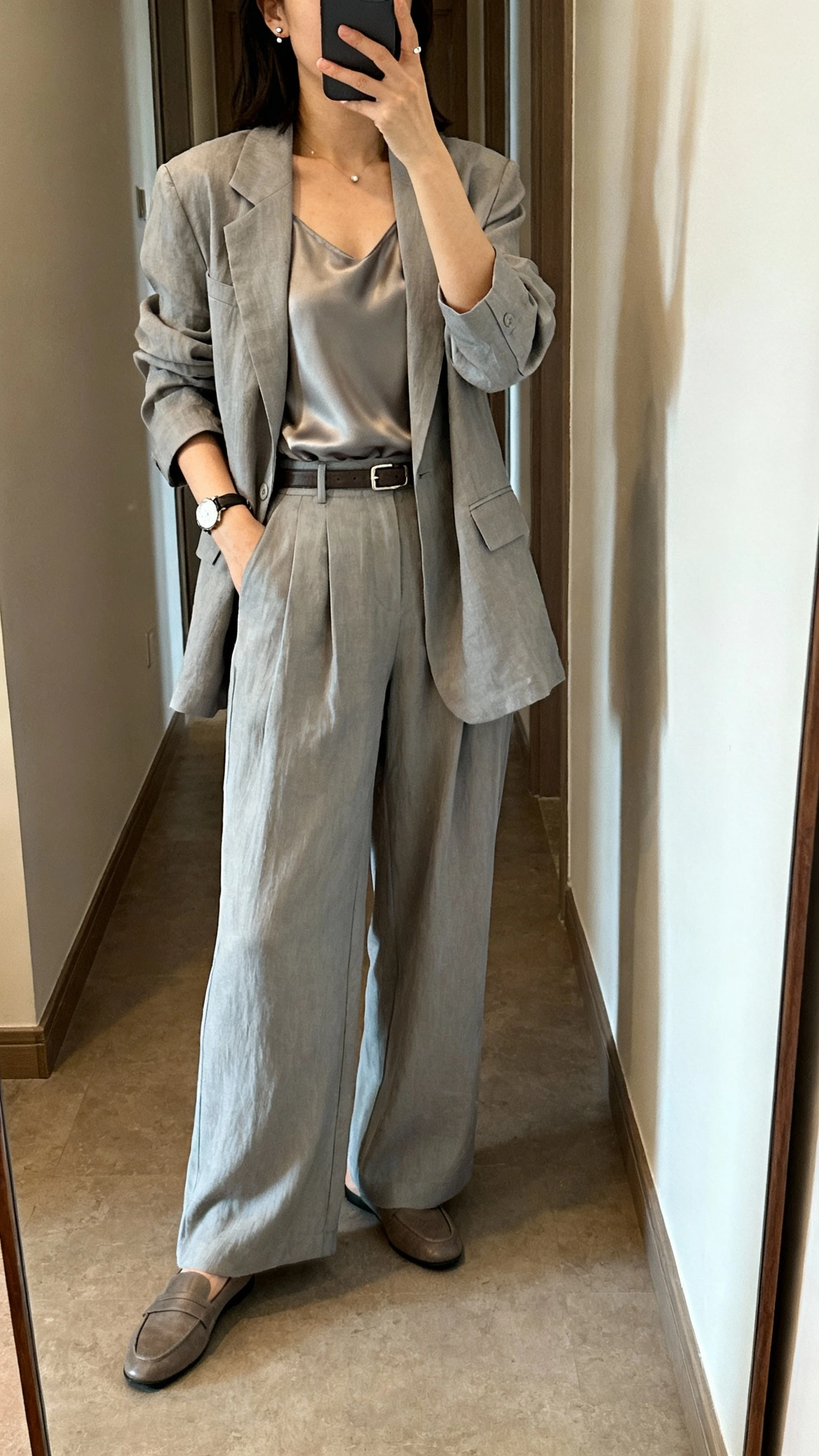 iPhone hallway mirror selfie of a woman wearing a soft gray relaxed blazer with coordinating wide-leg trousers, a silky shell top, low loafers, minimal watch and simple studs, slim belt; face not visible, natural indoor light, casual iPhone aesthetic.