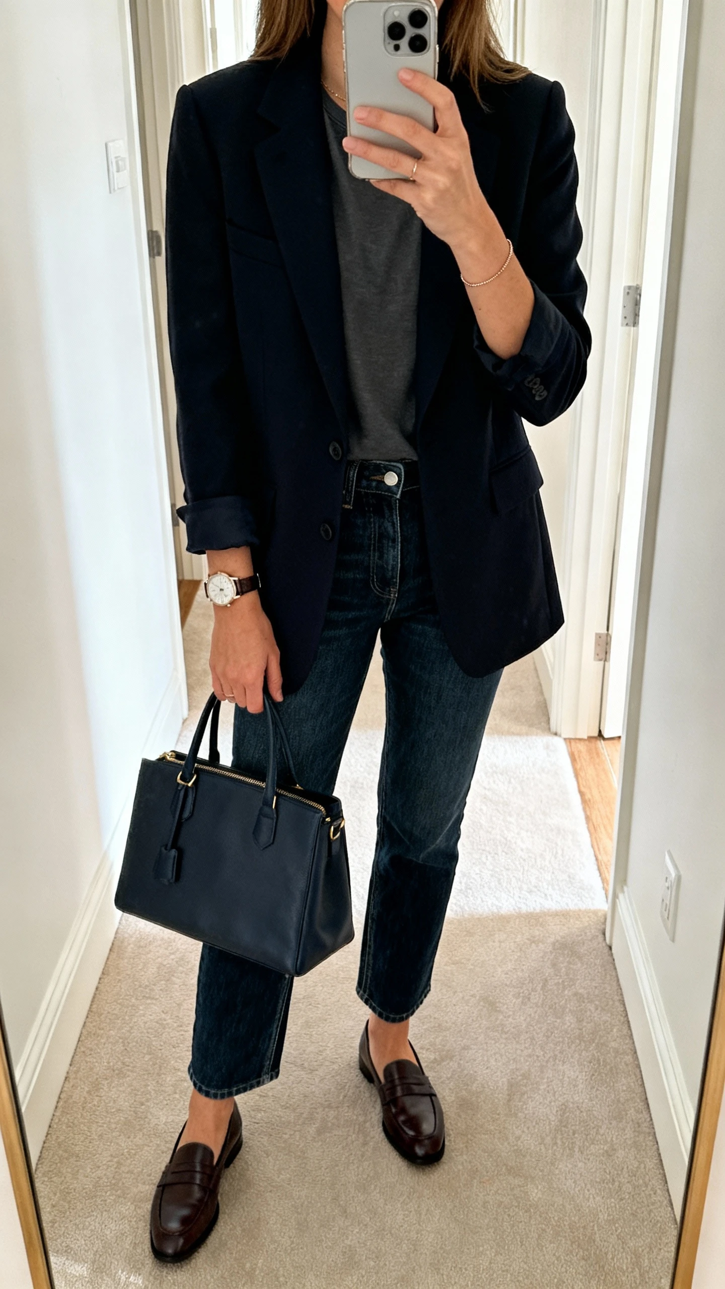 iPhone hallway mirror selfie of a woman wearing polished dark-wash denim, a tailored blazer over a tucked-in tee, loafers, structured tote, delicate watch, face not visible, morning light, casual iPhone aesthetic.