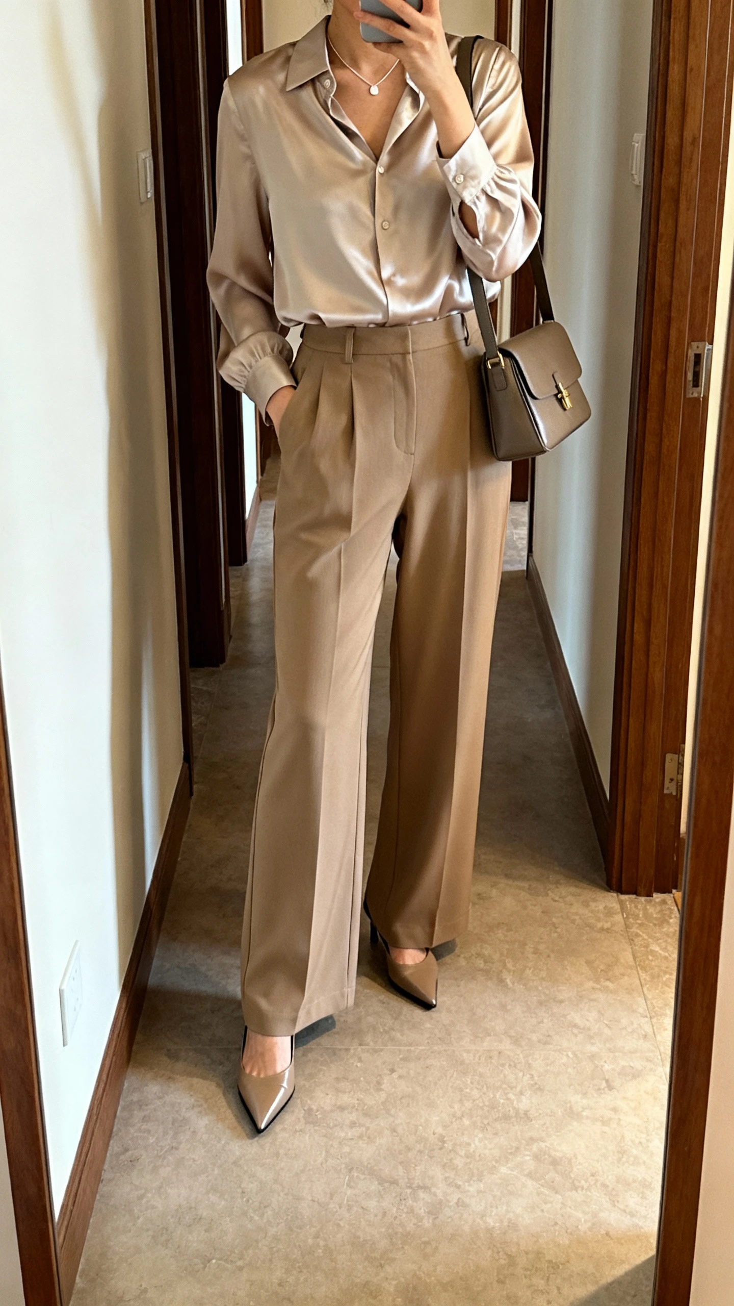 iPhone hallway mirror selfie of a woman wearing wide-leg trousers and a silk blouse, pointed-toe heels, minimalist necklace, structured shoulder bag, face not visible, warm indoor lighting, natural iPhone photo.