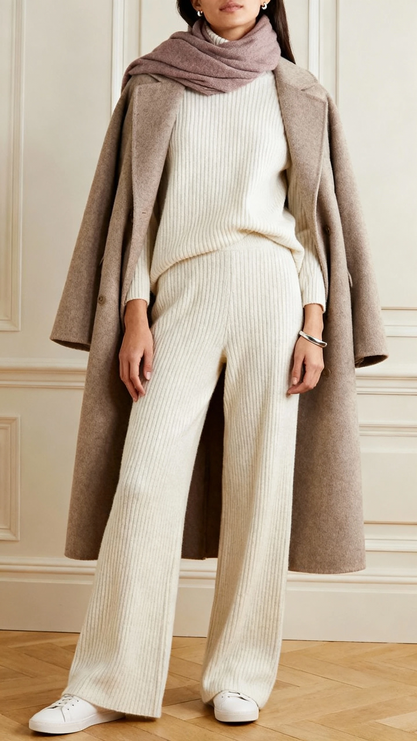 iPhone living-room mirror selfie of a woman in a monochrome rib-knit sweater with matching wide-leg knit pants, minimal white sneakers, a longline wool coat, a soft scarf in a slightly deeper tone, understated stud earrings and a sleek bangle; cropped at shoulders, face not visible, soft indoor light, natural iPhone photo quality.